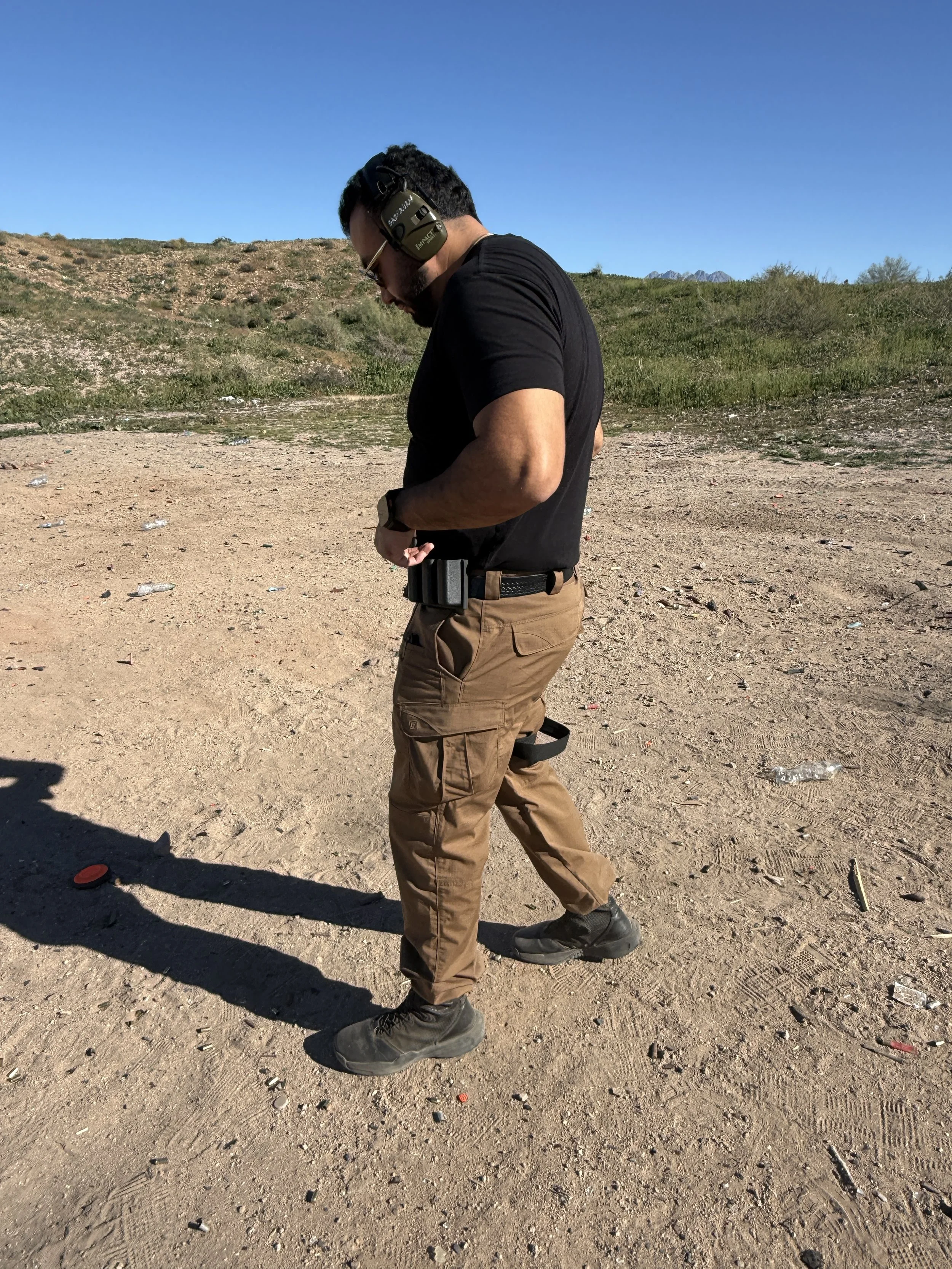 Man in black t-shirt and brown cargo pants standing outdoors on dirt ground, wearing hearing protection and looking down at his belt.