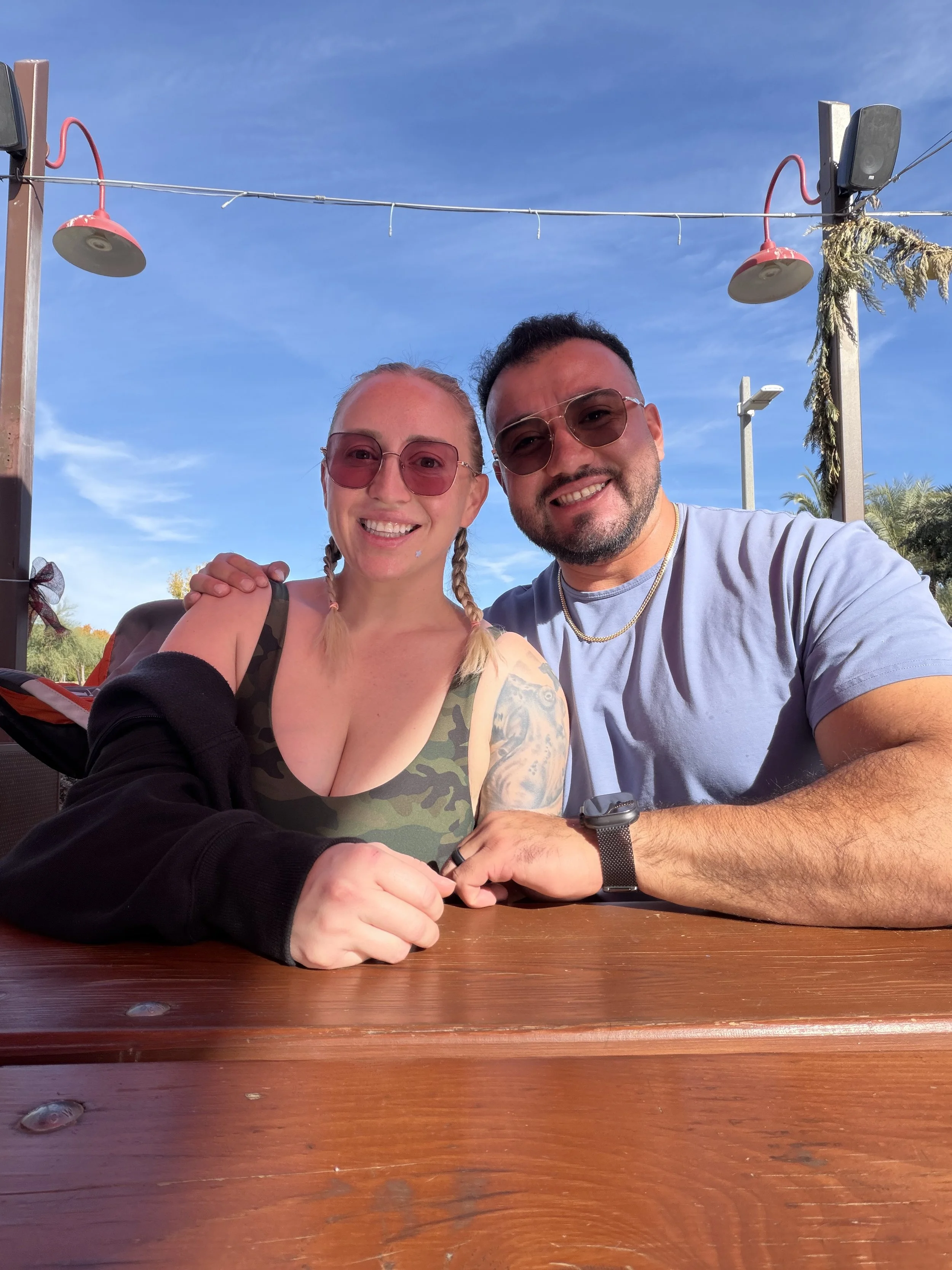 A smiling man and woman sitting together at a wooden table outdoors, both wearing sunglasses under a blue sky with string lights and outdoor lights visible overhead.