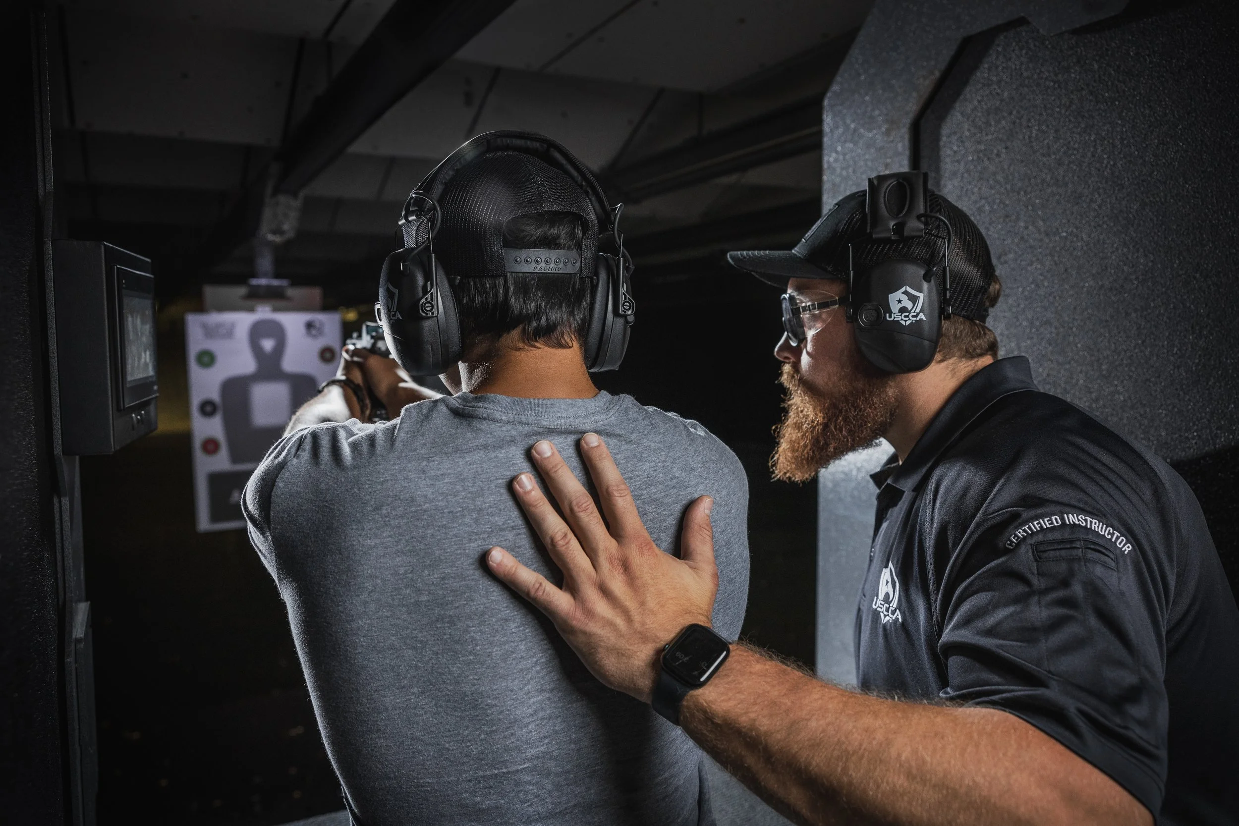 A firearms training instructor coaching a student at a shooting range, with both wearing protective ear gear and the instructor providing guidance.
