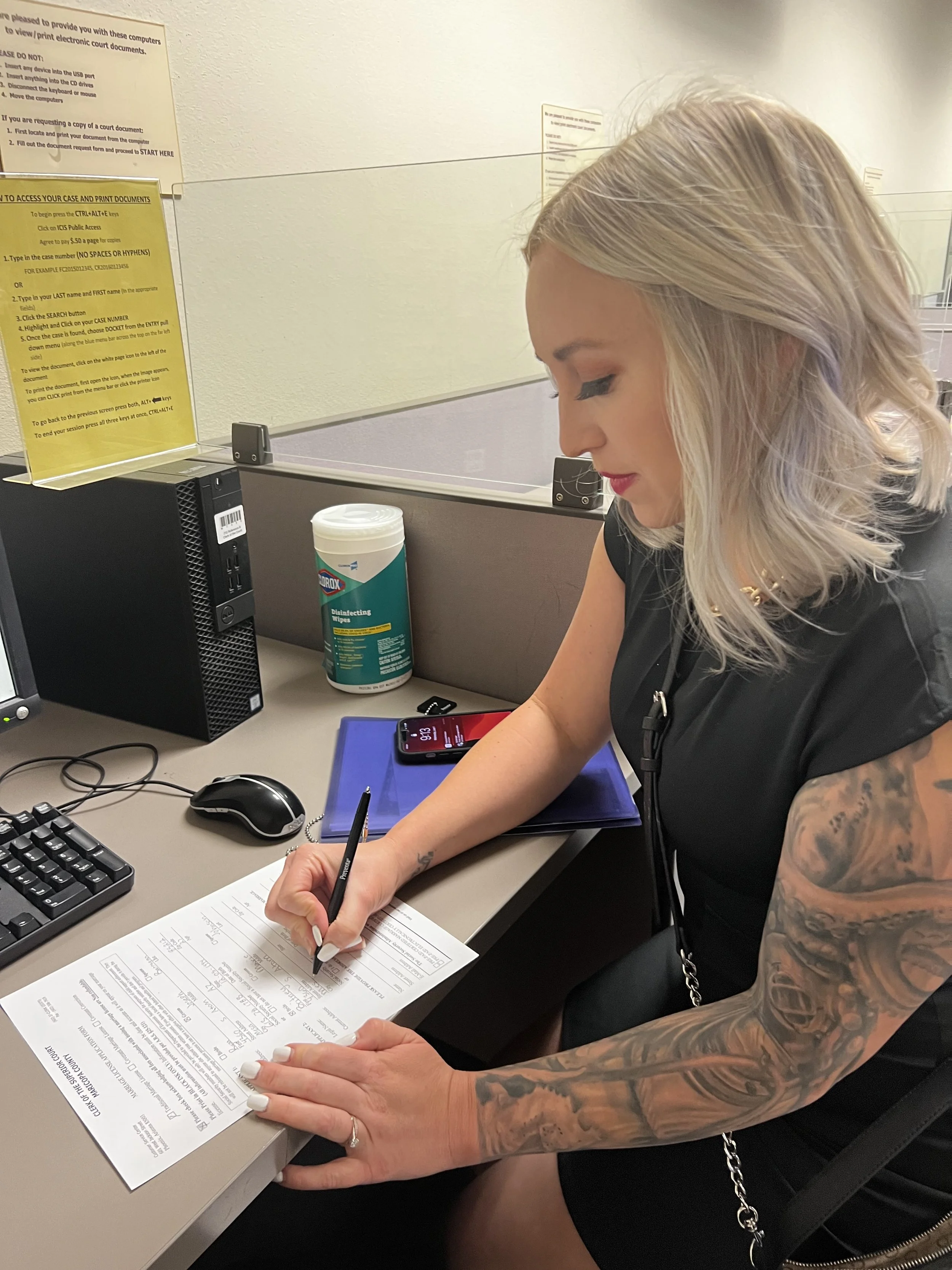 A woman with blonde hair and tattoos on her arm sitting at a desk, filling out a form with a black pen. On the desk, there is a computer mouse, a keyboard, a smartphone, a blue folder, disinfectant wipes, and a desktop computer. Behind her, there is a yellow informational sign on the wall.