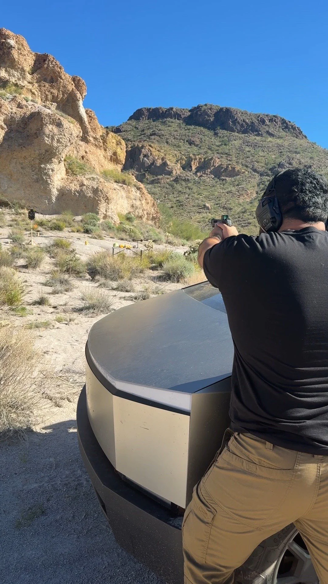 A person wearing a black shirt and khaki pants shooting a firearm at a shooting range with rocky desert landscape and blue sky in the background.