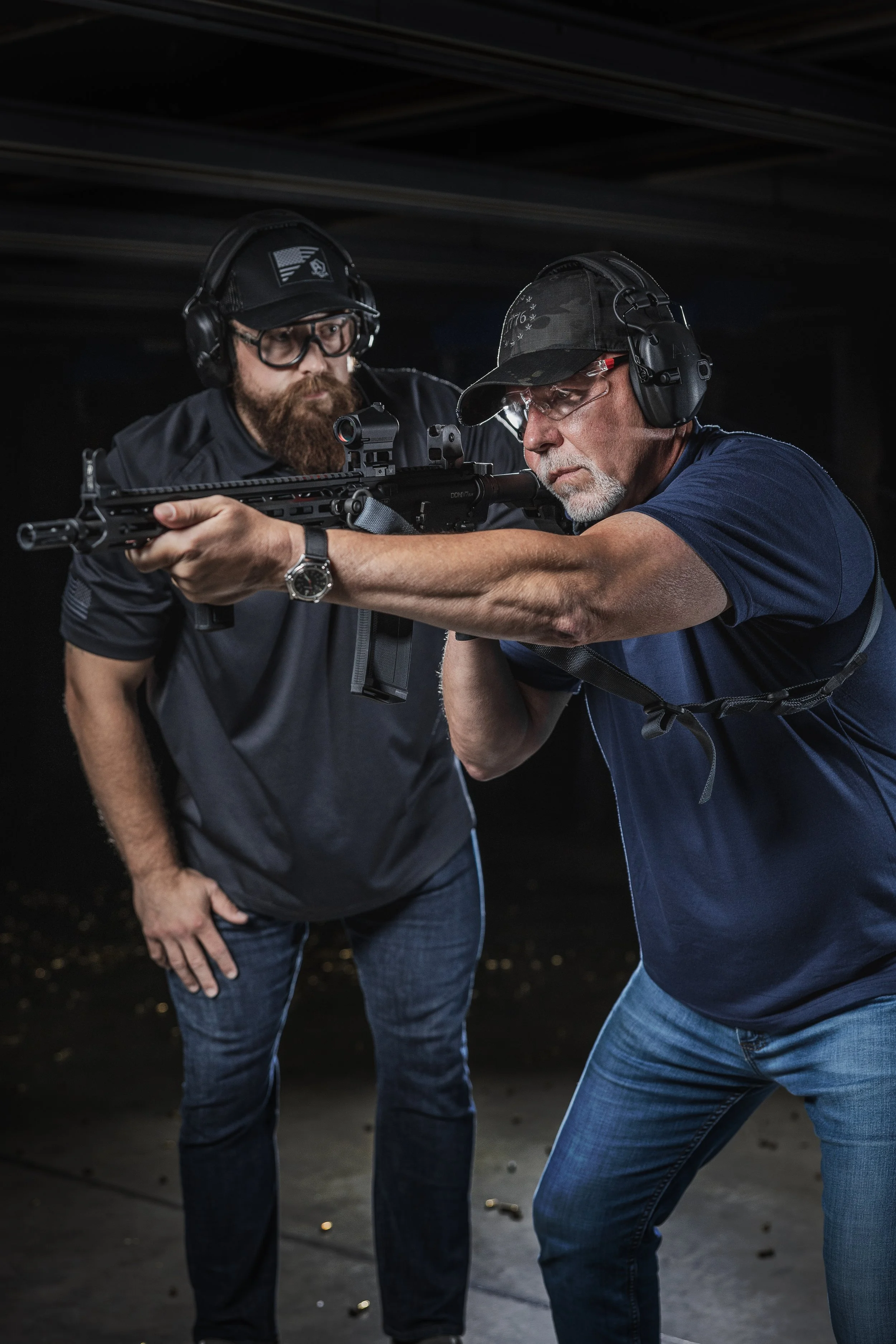 Two men at a shooting range practicing firearm safety, one instructing the other with a rifle.