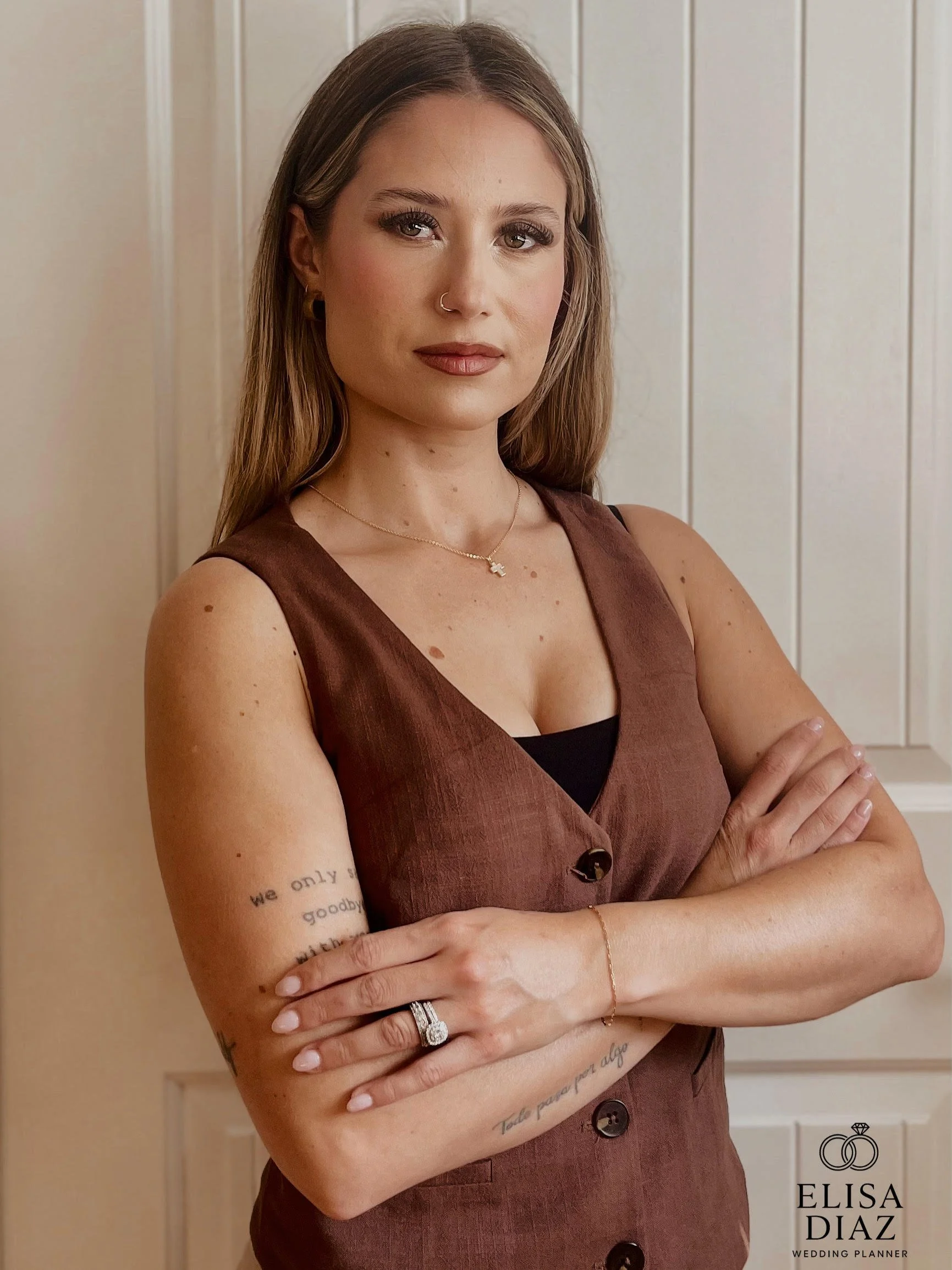 A woman with long, light brown hair, wearing a sleeveless brown dress with a black top underneath, stands against a beige wall with vertical paneling, with arms crossed.