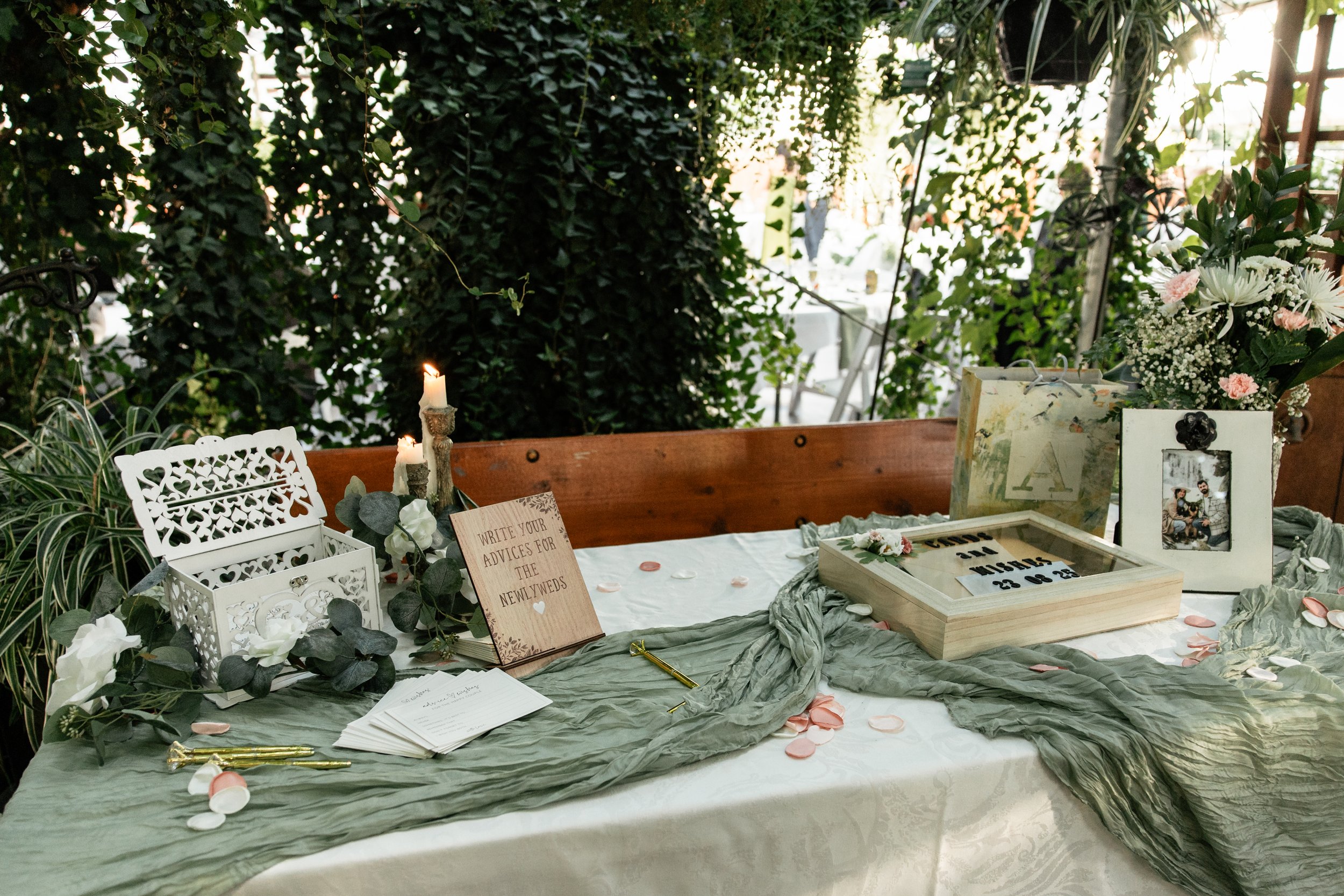 A decorated wedding guestbook table with candles, flowers, a sign for writing advice, and photo frames, set outdoors with lush greenery in the background.
