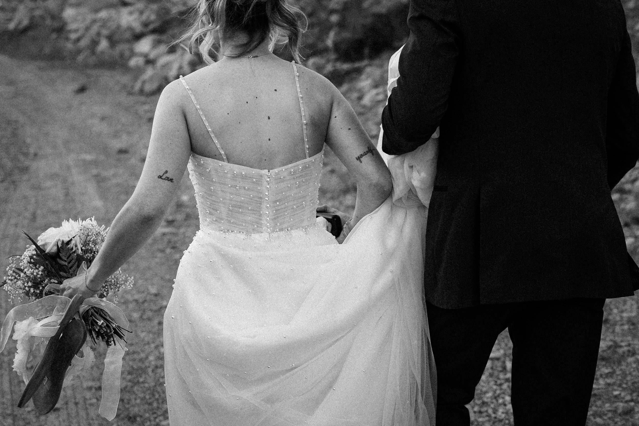 A black and white photo of a bride holding a bouquet walking arm in arm with a man, outdoors on a dirt path.