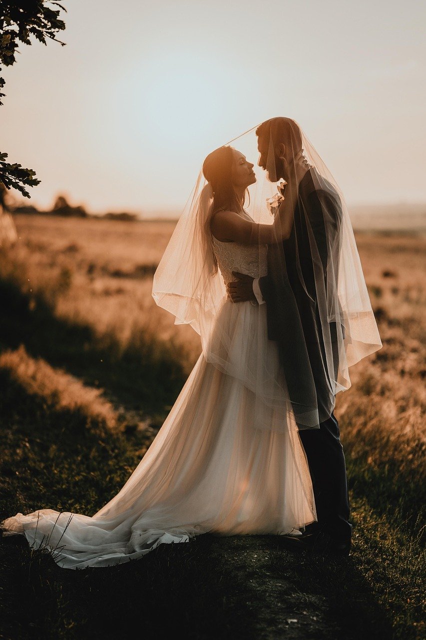 A bride and groom standing close together outdoors during sunset, under a veil, with the bride touching the groom's face, overlooking a grassy field in the background.