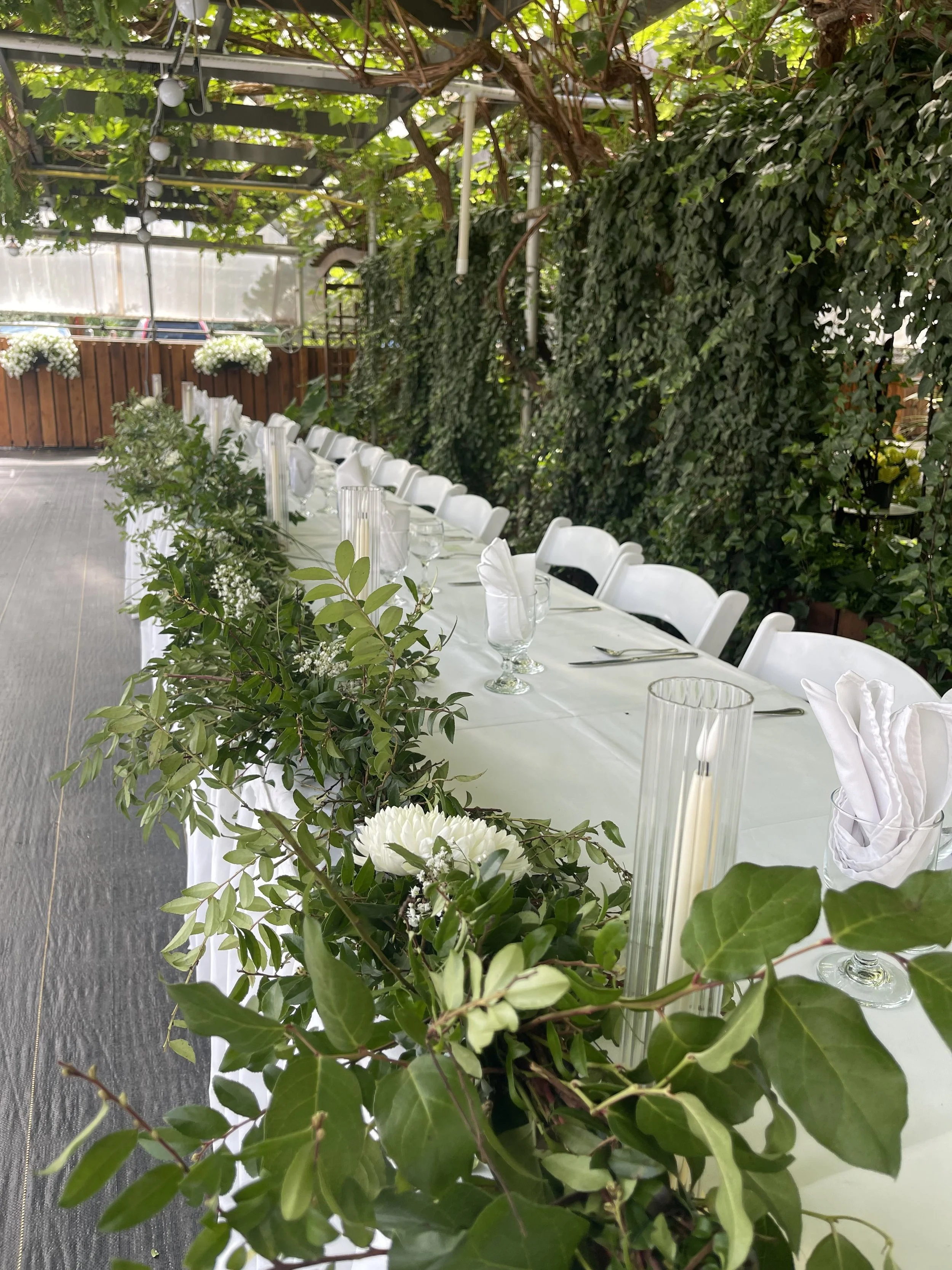 Wedding or event table set outdoors with white chairs, white tablecloth, glassware, silverware, and napkins, decorated with green foliage and white flowers, under a trellis with green vines.
