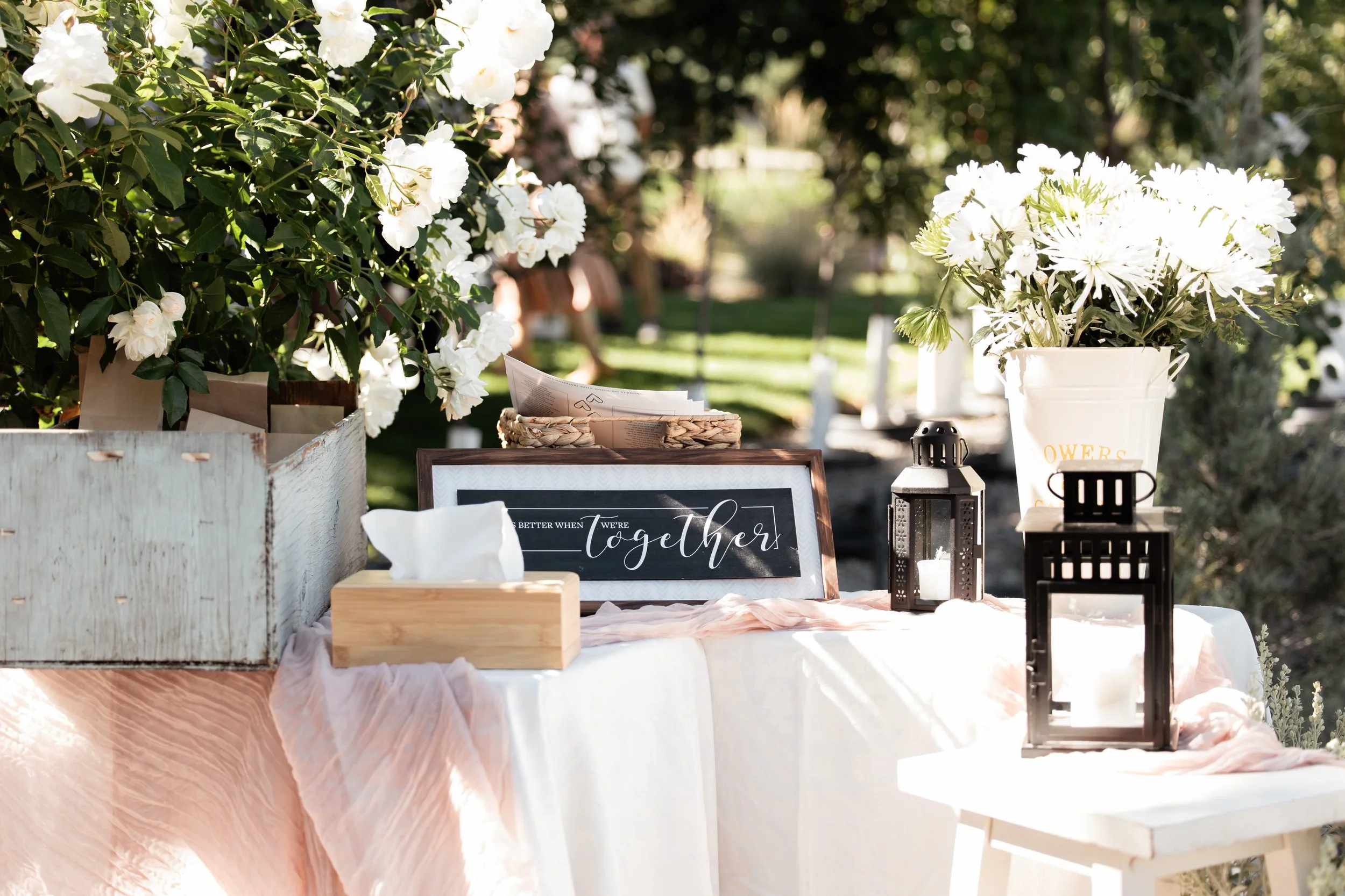 An outdoor table decorated with white flowers, candles, a tissue box, and a framed sign that reads 'Better when we're together,' set in a garden with green trees and sunlight.