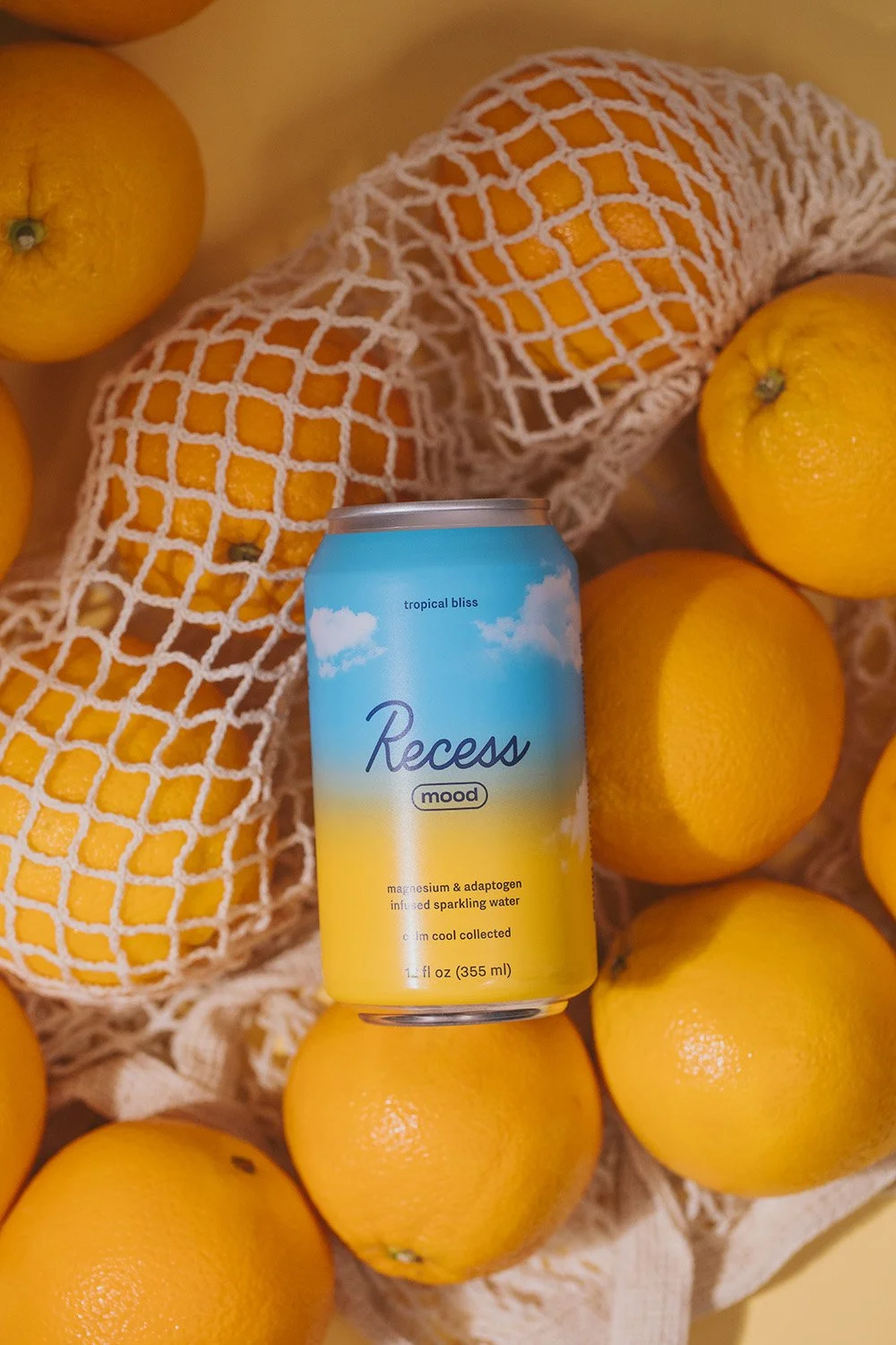 Lifestyle product photography of Recess Mood tropical bliss sparkling water can nestled among fresh oranges in cotton mesh produce bag