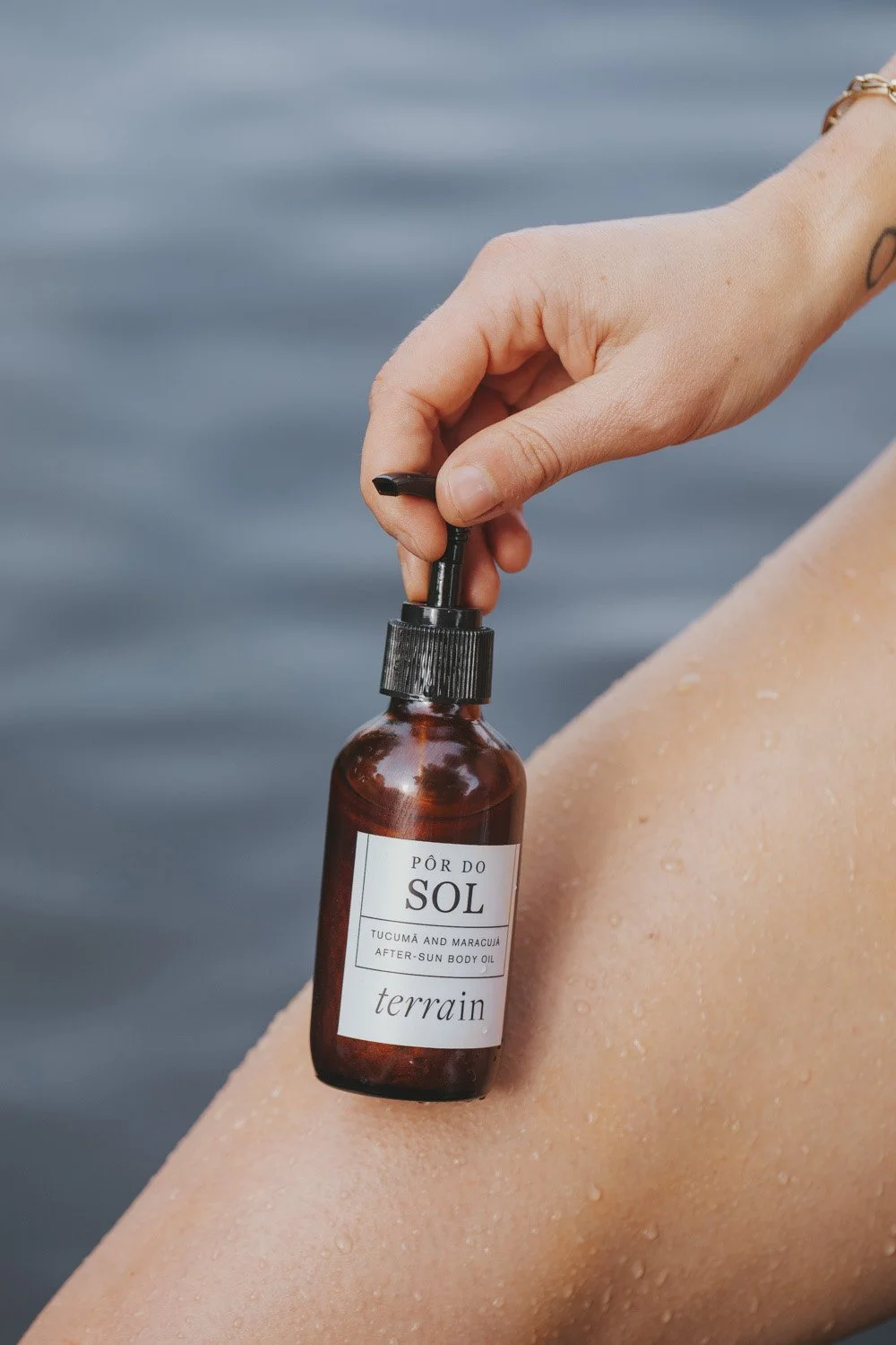 Hand holding amber glass bottle of Pôr do Sol after-sun body oil by Terrain against blurred water backdrop