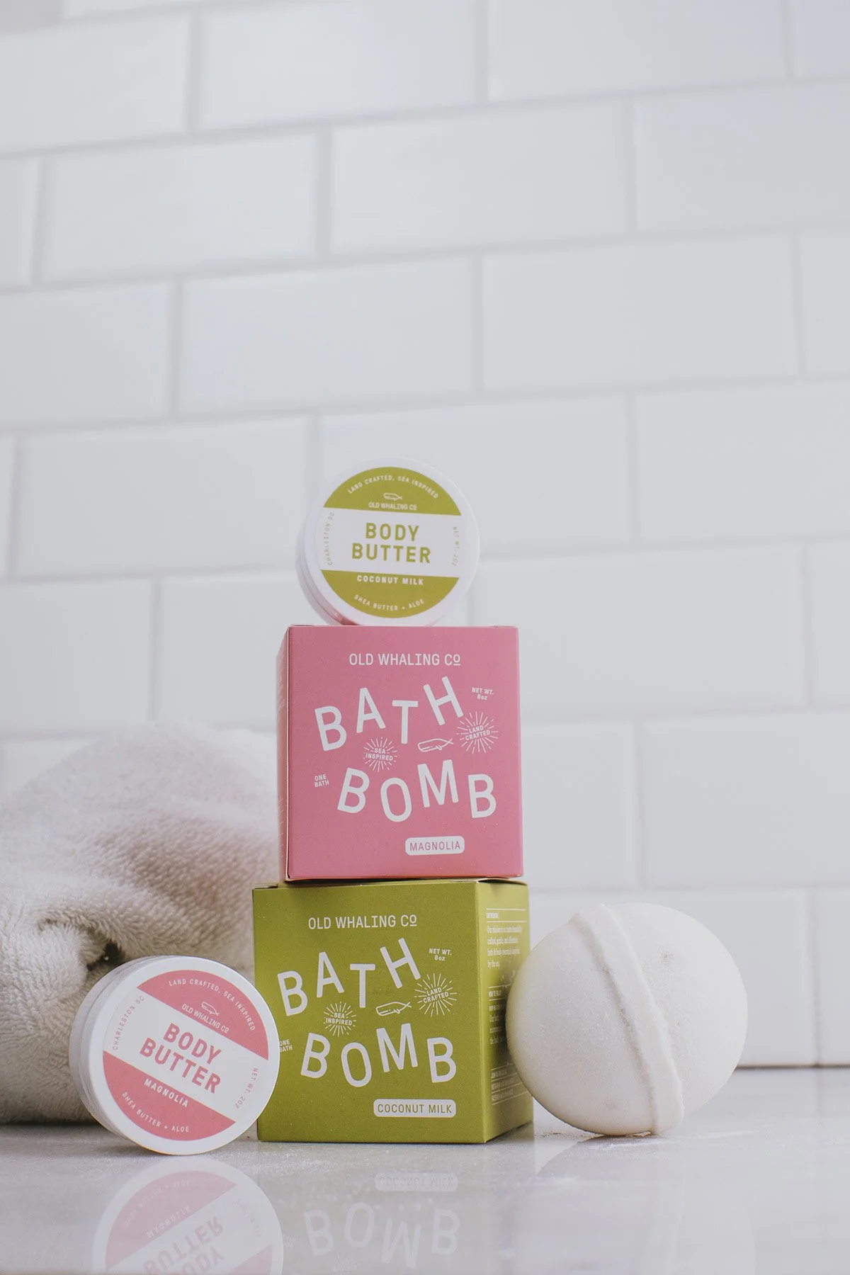 Old Whaling Co bath bomb boxes in pink magnolia and coconut milk scents stacked with body butter tins and white towel against white subway tile
