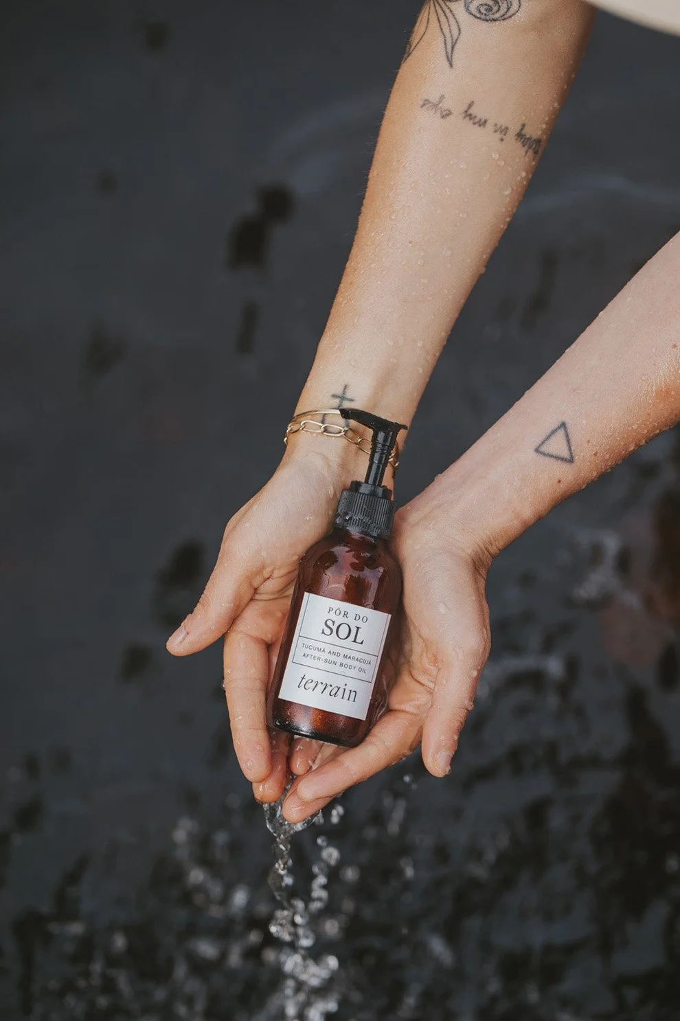 Hands holding Por do Sol after sun body oil bottle by Terrain with water droplets, person wearing gold chain bracelet and visible tattoos against dark background