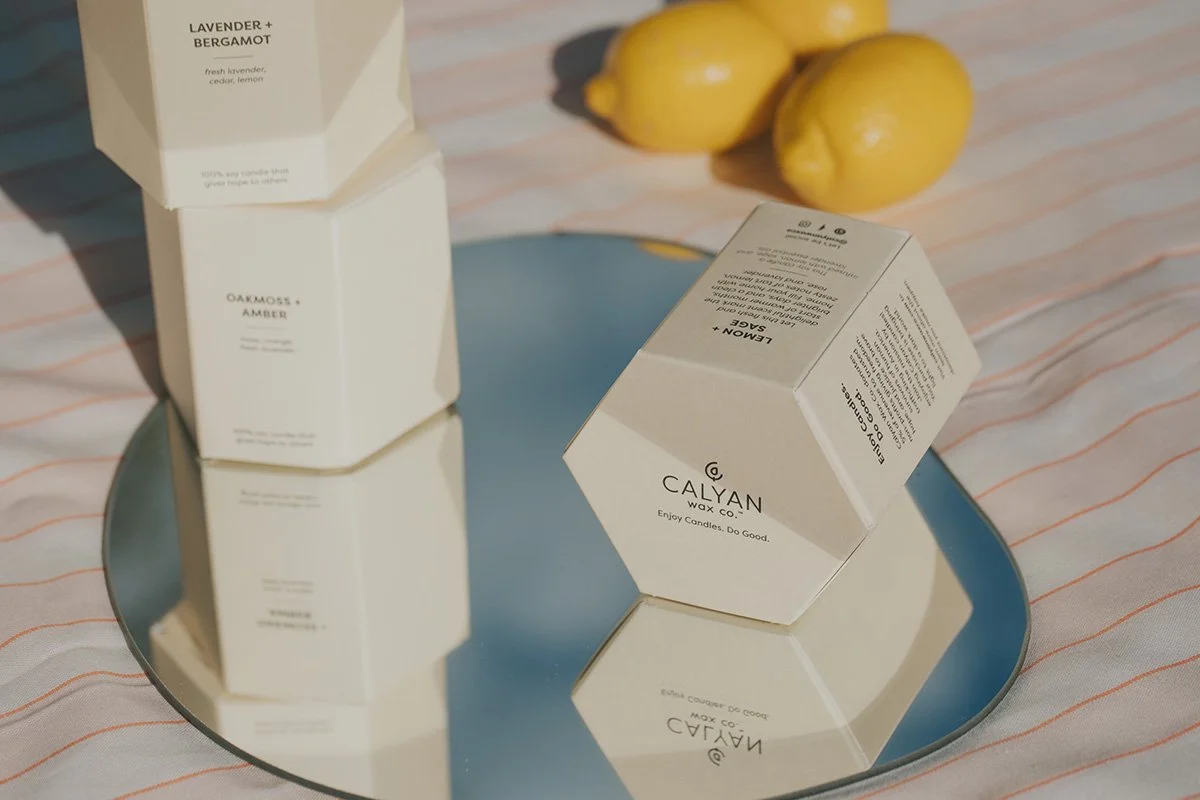 Styled product flat lay featuring Calyan Wax Co. hexagonal candle packaging with lavender bergamot and oakmoss amber scents on teal plate with fresh lemons