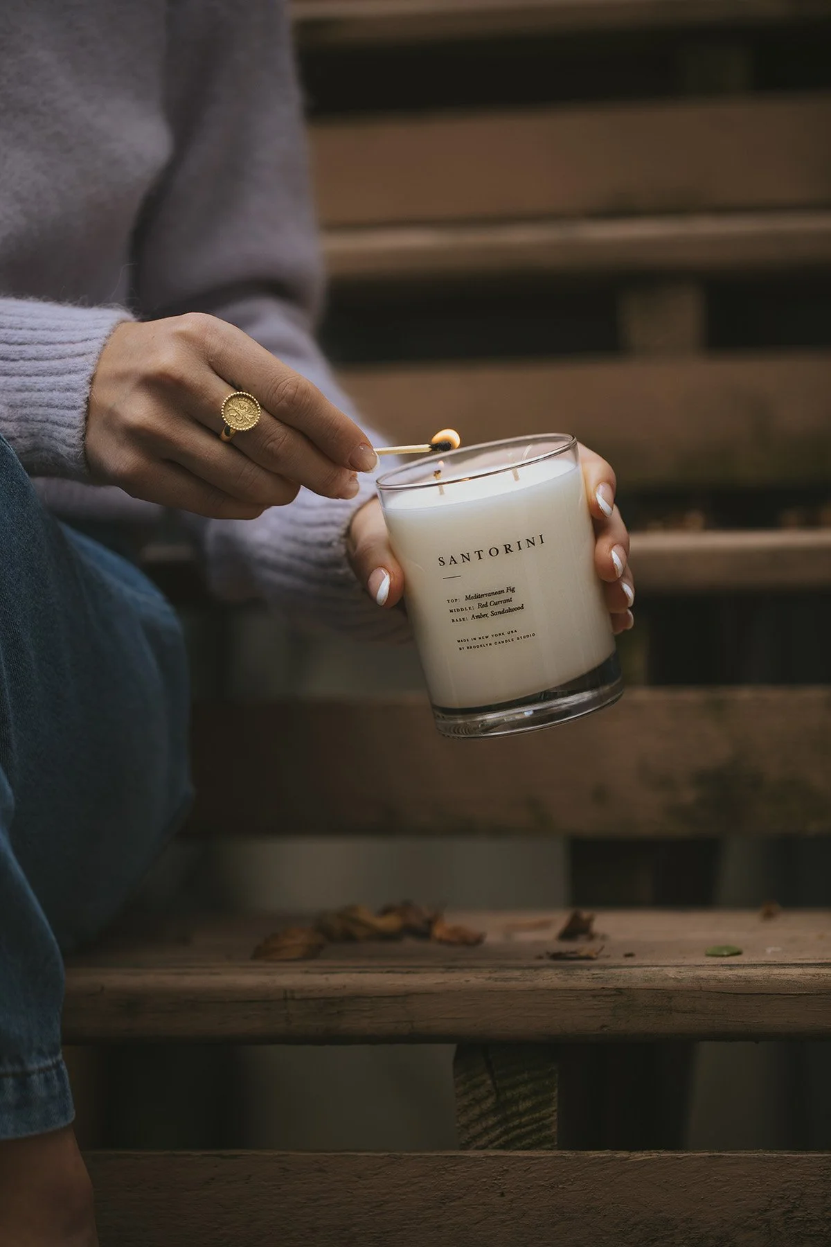 Hands wearing gold ring lighting Santorini candle with match, person in gray sweater sitting by fireplace in cozy setting