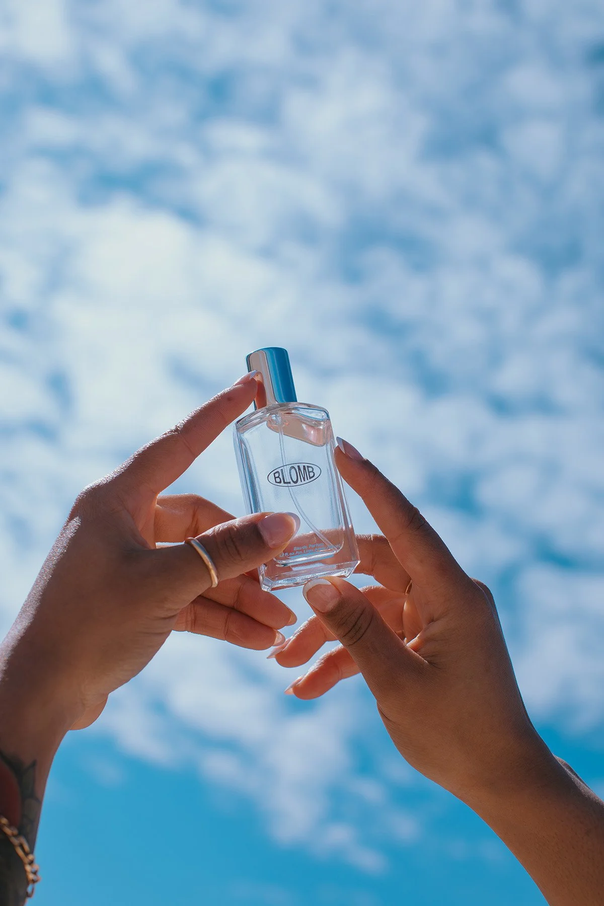 Hands holding clear Blomb perfume bottle with blue cap against bright blue sky with white clouds