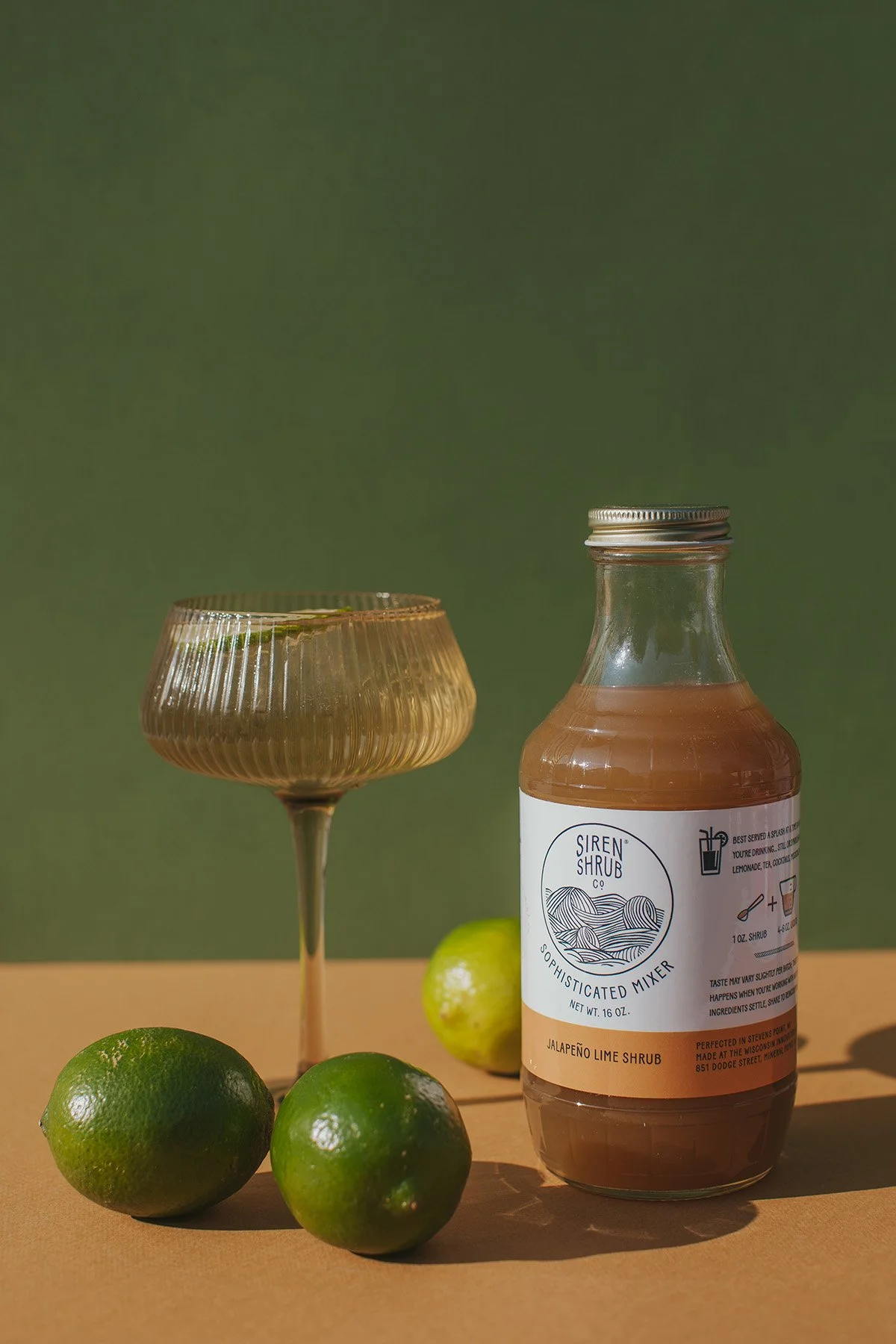 Siren Shrub Co jalapeño lime sophisticated mixer bottle with fresh limes and vintage coupe glass on wood surface against sage green background