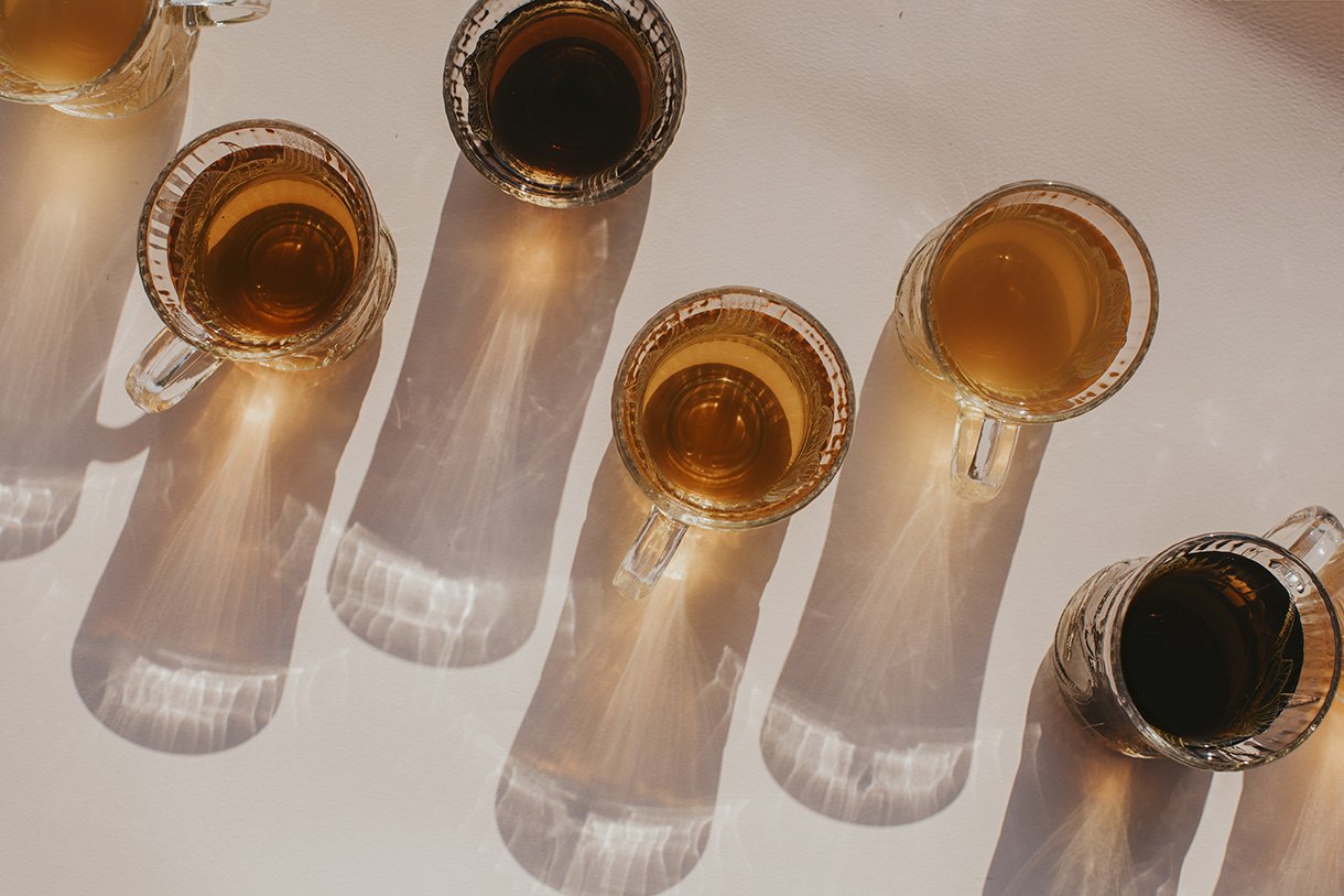 Six glasses of various beverages with different shades, casting long shadows on a white surface.