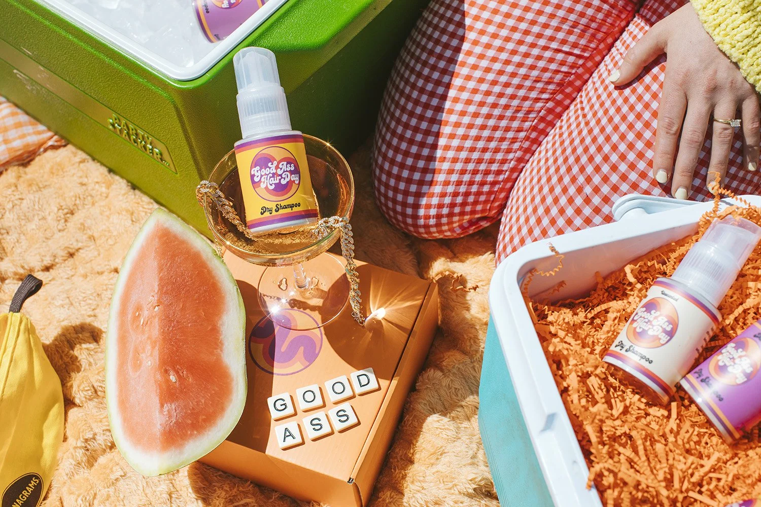 A watermelon slice, a small bottle of dry shampoo labeled 'Good Ass Hair Day,' a glass with a booklet spelling 'GOOD ASS,' and a box with orange shredded paper containing additional bottles of dry shampoo. The scene is set on a orange textured blanket, with a high angle view showing a person's hand with a ring and checkered orange and white pants.