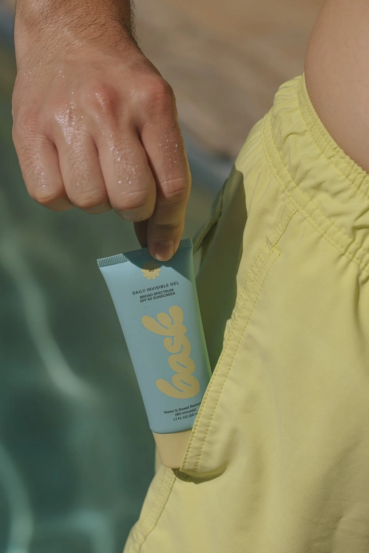 Hand holding Bask daily invisible gel SPF 40 broad spectrum sunscreen in yellow swim shorts pocket with water droplets on skin
