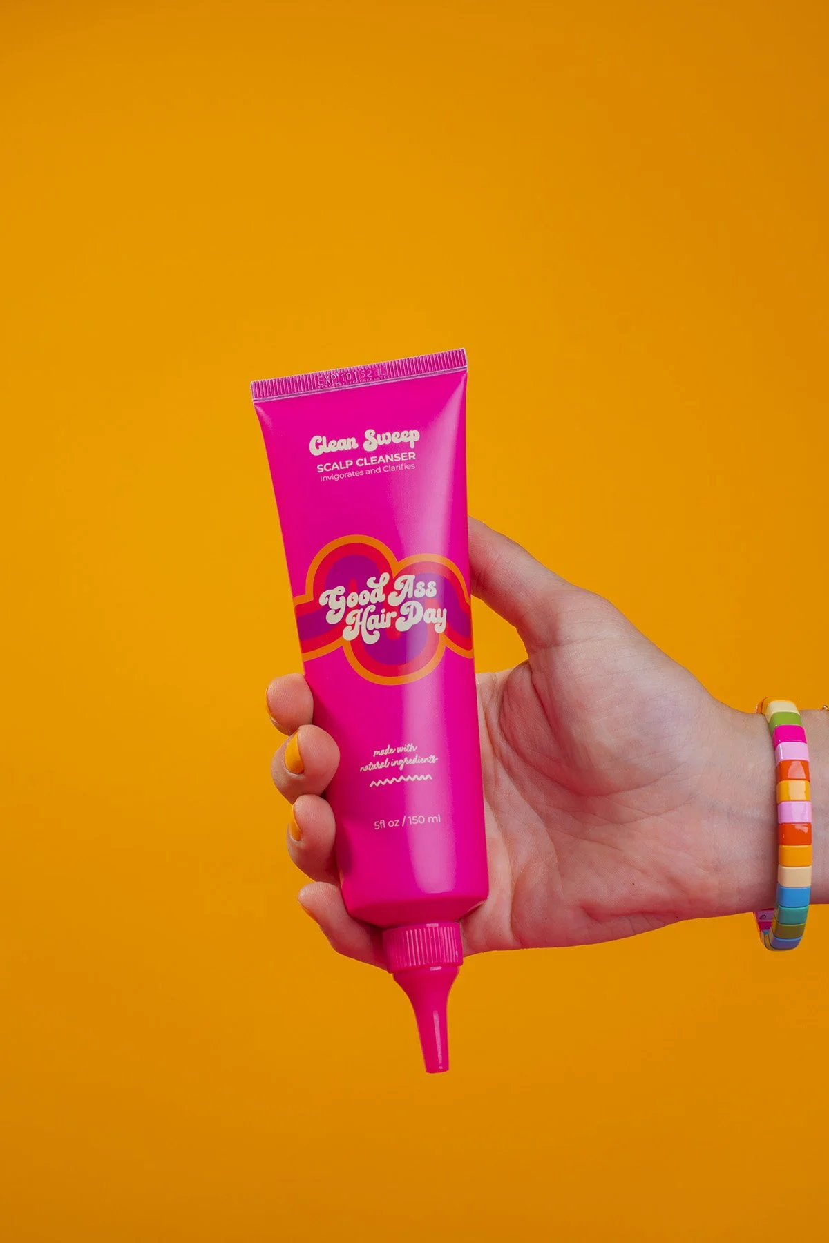 Hand with colorful beaded bracelet holding bright pink Good Ass Hair Day Clean Sweep scalp cleanser tube against vibrant orange background