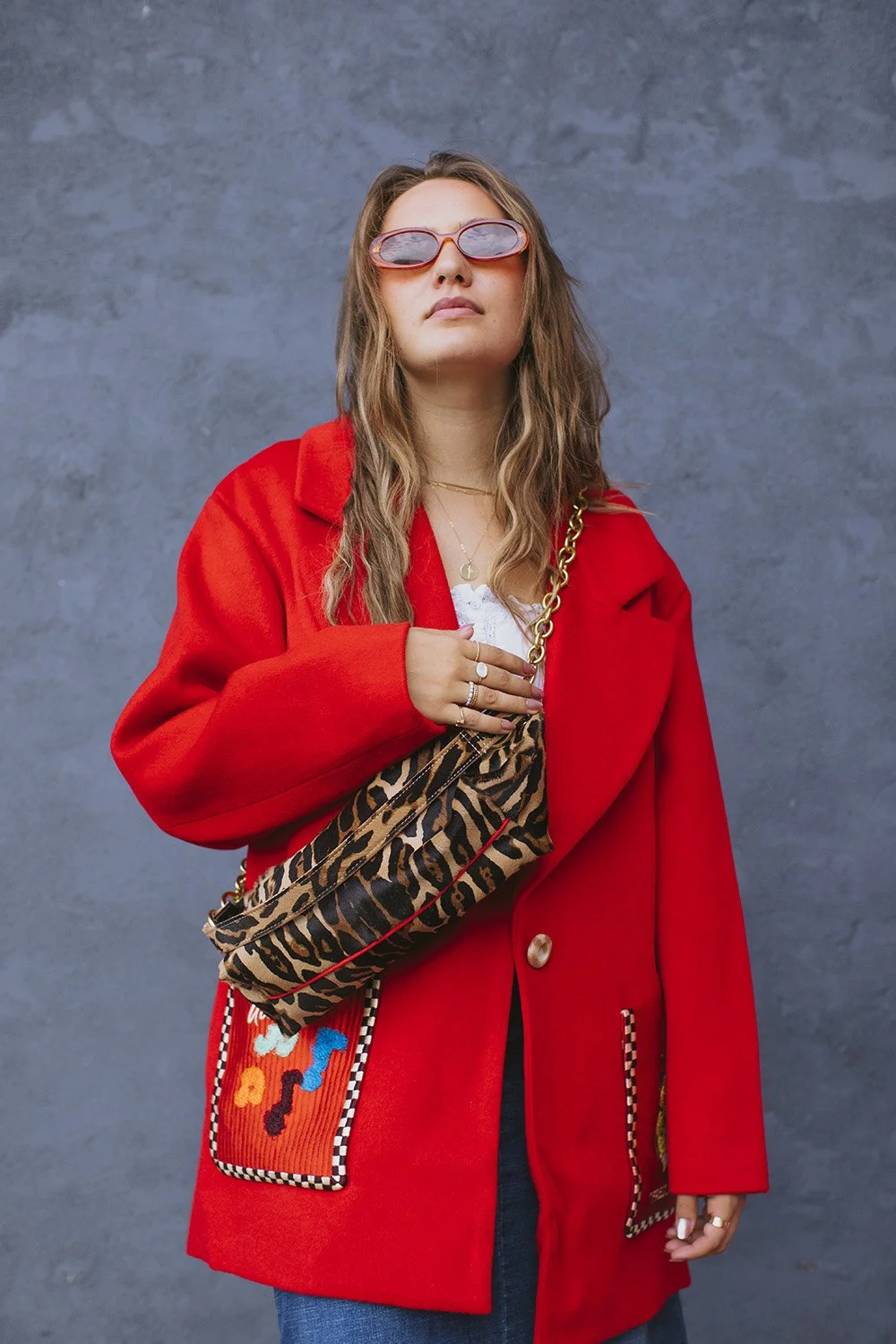 Fashion portrait of woman in oversized red blazer with embroidered pockets wearing purple-tinted sunglasses and animal print crossbody bag against textured gray wall