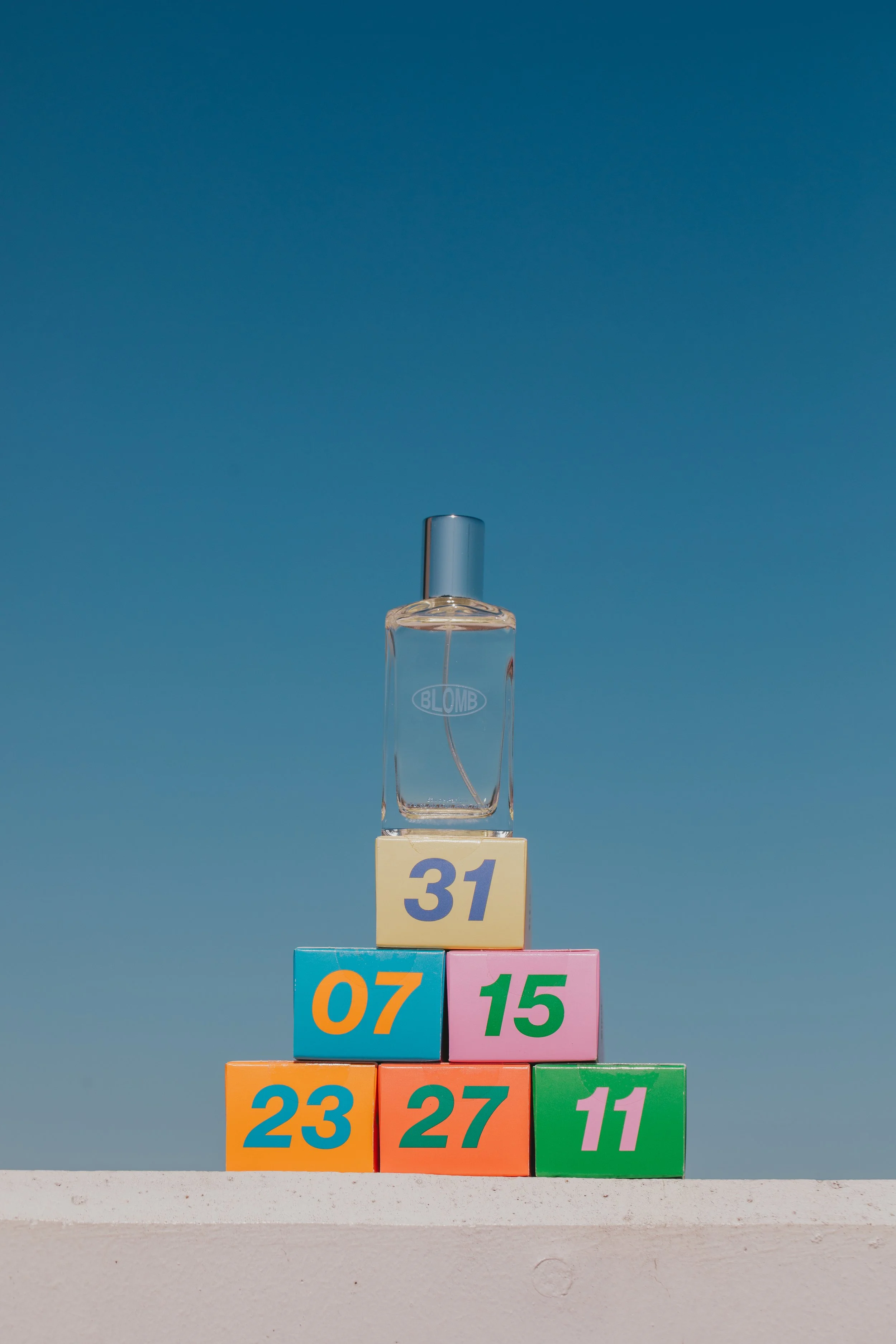 Colorful blocks with numbers stacked in a pyramid, topped with a transparent perfume bottle, against a blue sky background.