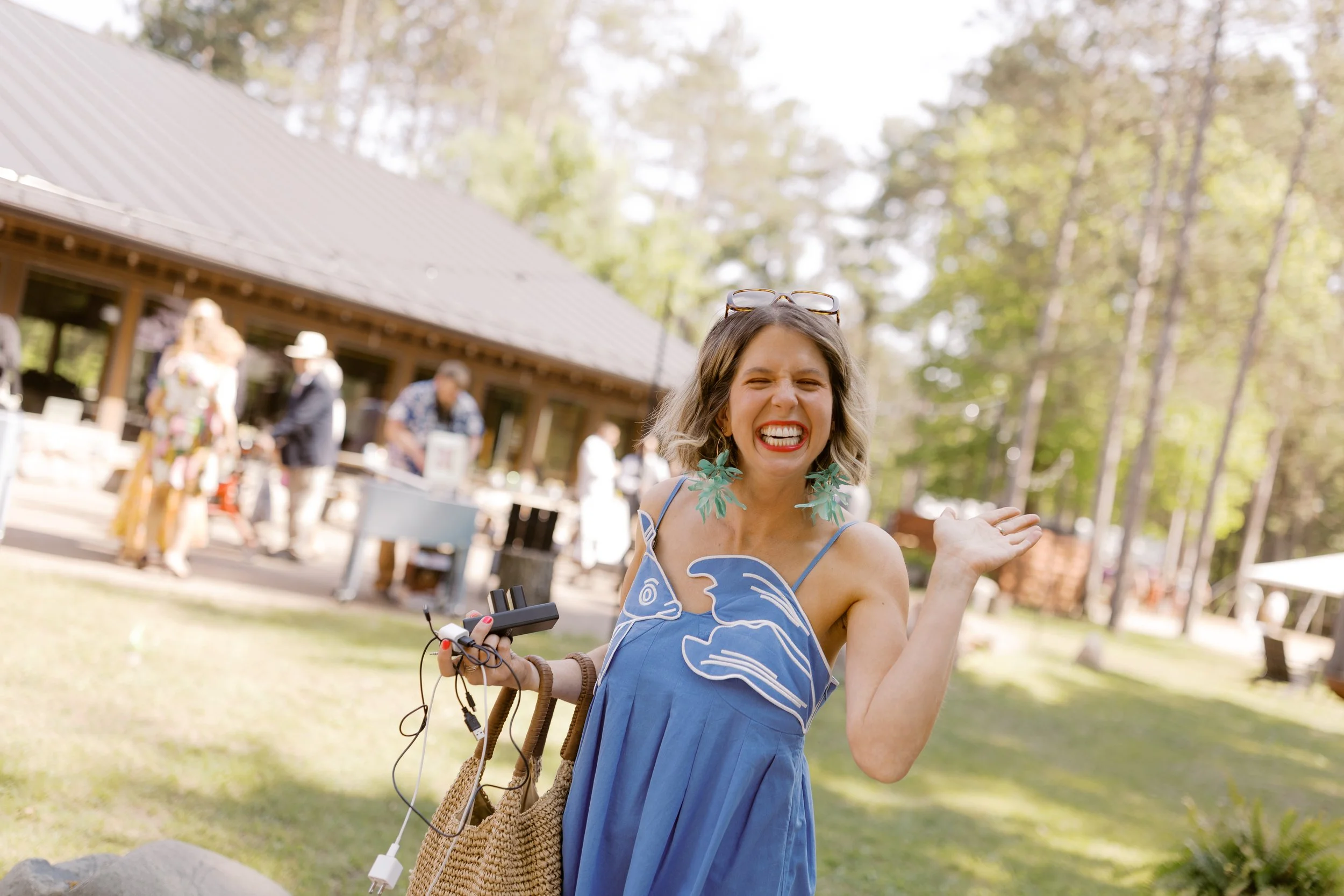 Joyful woman in blue sundress with palm leaf earrings laughing at outdoor summer event with rustic pavilion in background