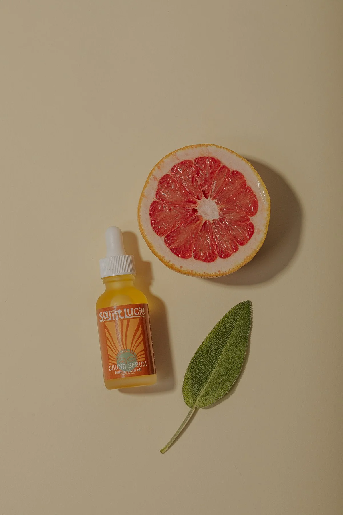Saint Lucie sauna serum bottle with orange sunburst design beside pink grapefruit slice and sage leaf on neutral beige background