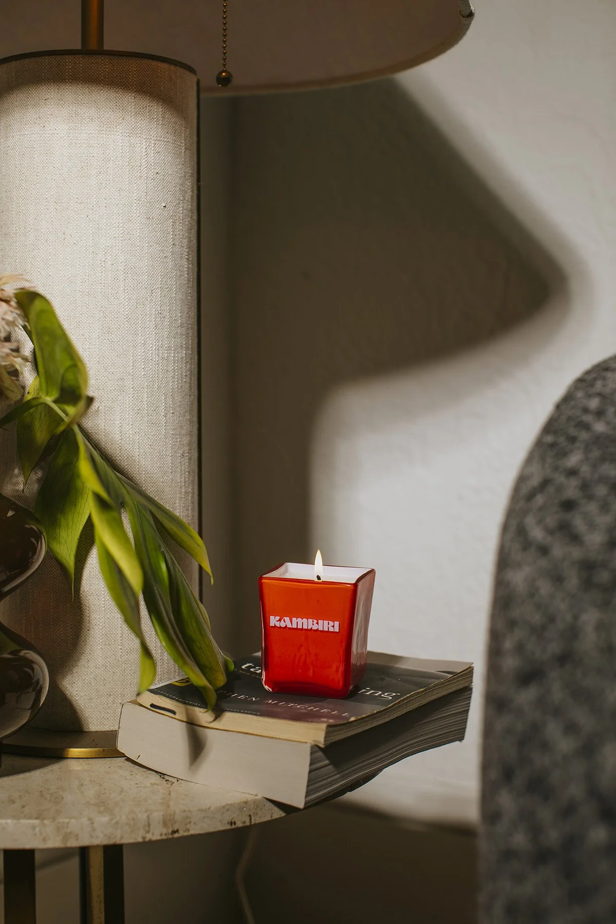 Red Kambini candle burning on stack of books next to plant and lamp with natural lighting and cozy home decor styling