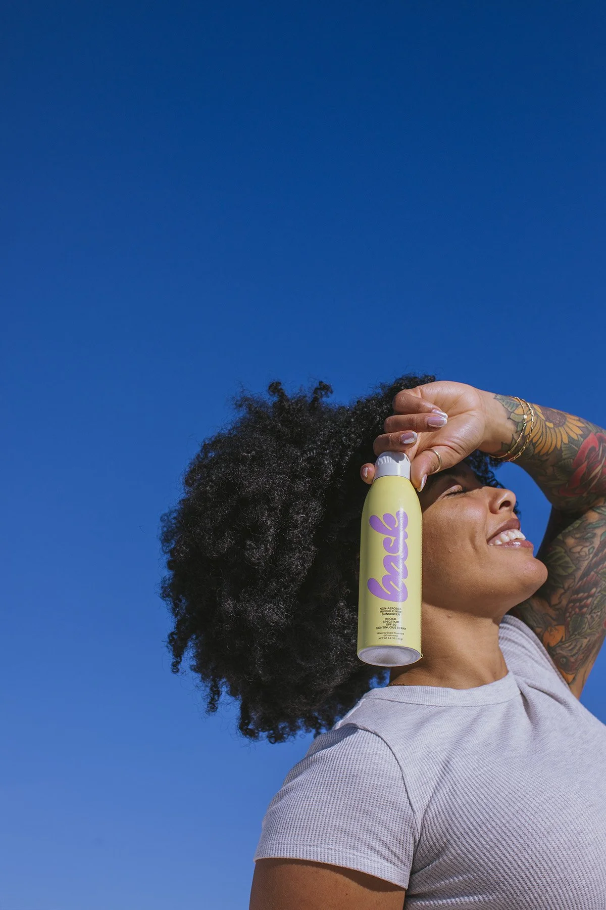 Low-angle lifestyle shot of woman with natural afro hair spraying Bask Ireland yellow aerosol product against bright blue sky