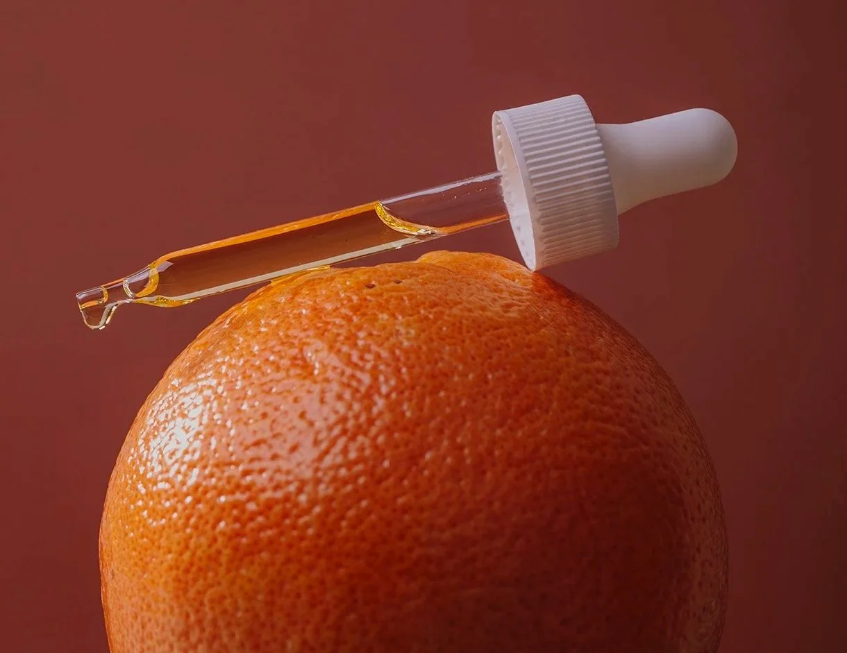 Minimalist product photography of glass dropper with golden serum resting on fresh orange against terracotta background