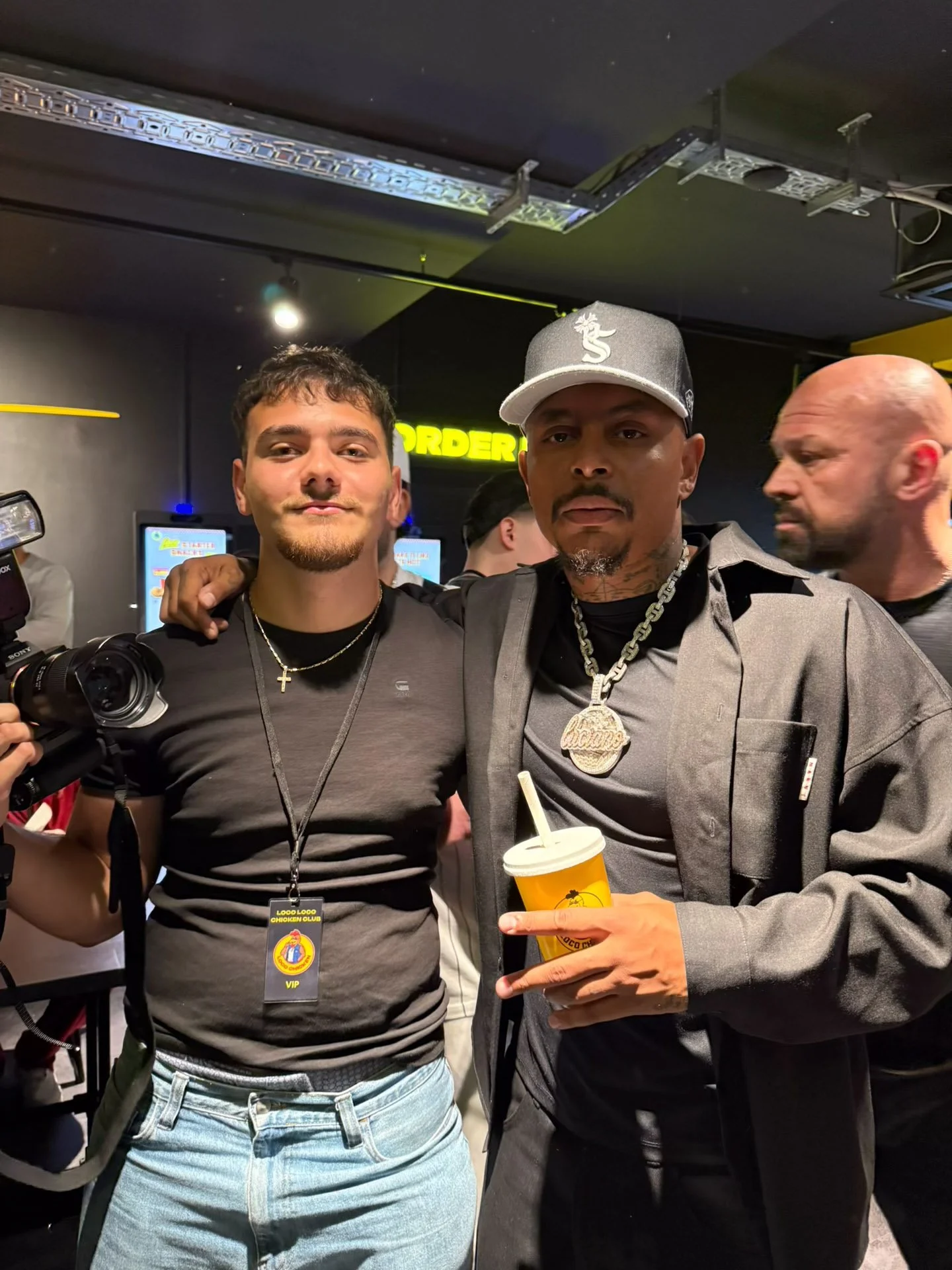 Zwei Männer stehen nebeneinander, die rechte Hand des Mannes auf der linken Schulter des anderen. Der Mann auf der linken trägt ein schwarzes T-Shirt, eine Halskette mit einem Kreuz und einen VIP-Ausweis. Der Mann auf der rechten trägt eine graue Jac