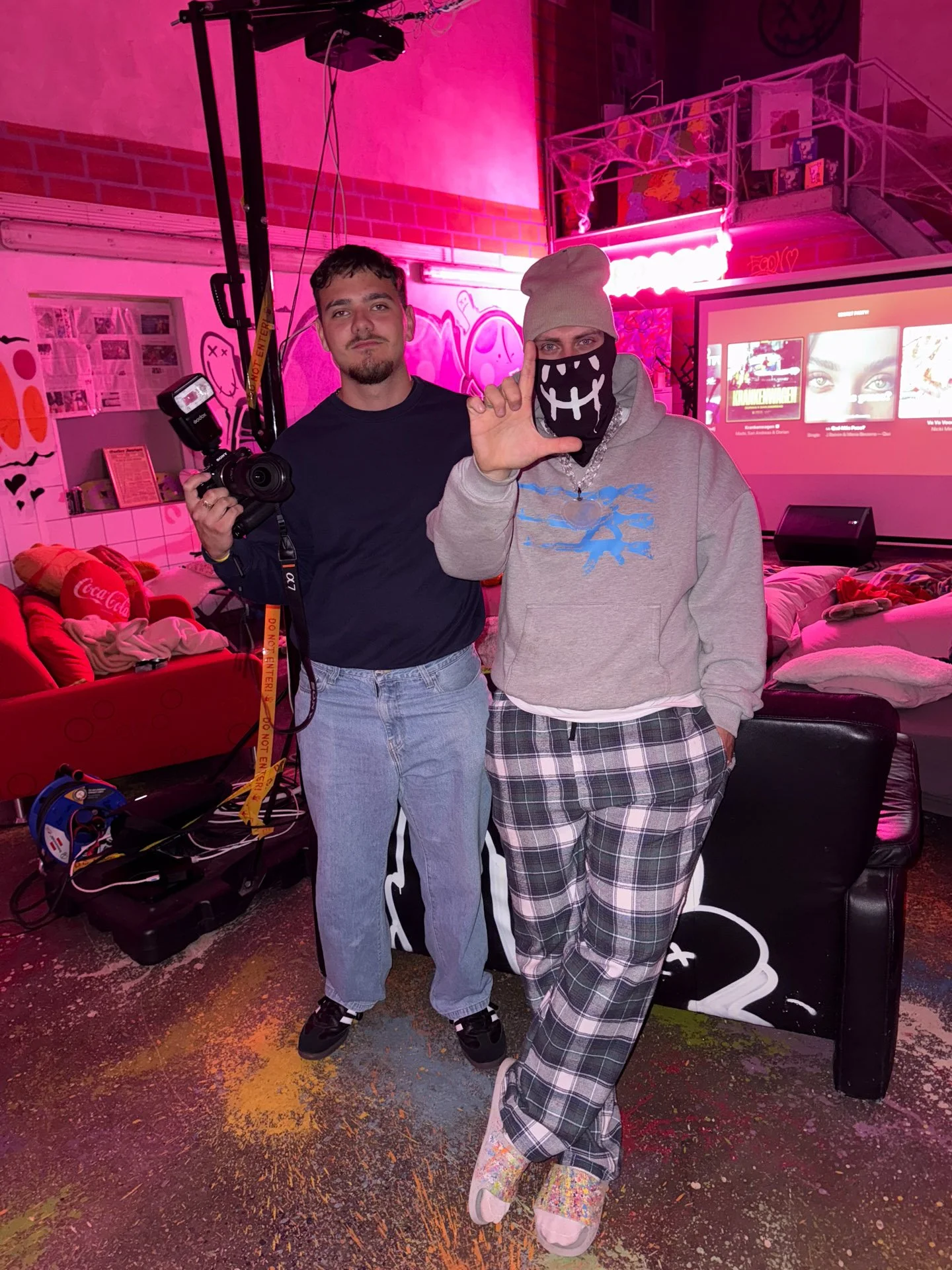 Sandro und Twentyoneseason, einer mit Kamera, stehen vor einem pink beleuchteten Hintergrund in einem Raum mit Graffitis und einem Fernseher.