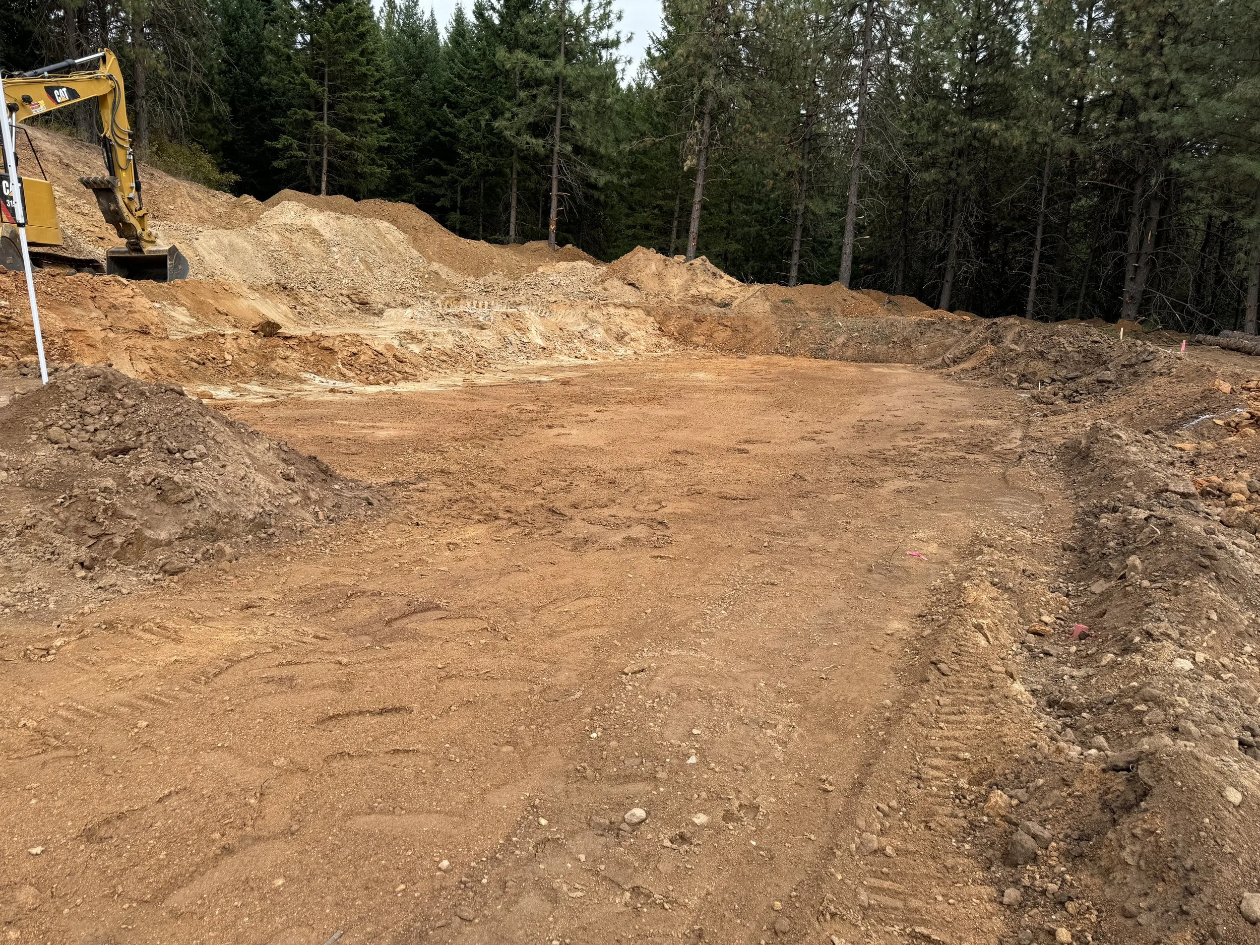 Construction site with a cleared dirt area, excavator in the background, surrounded by trees.