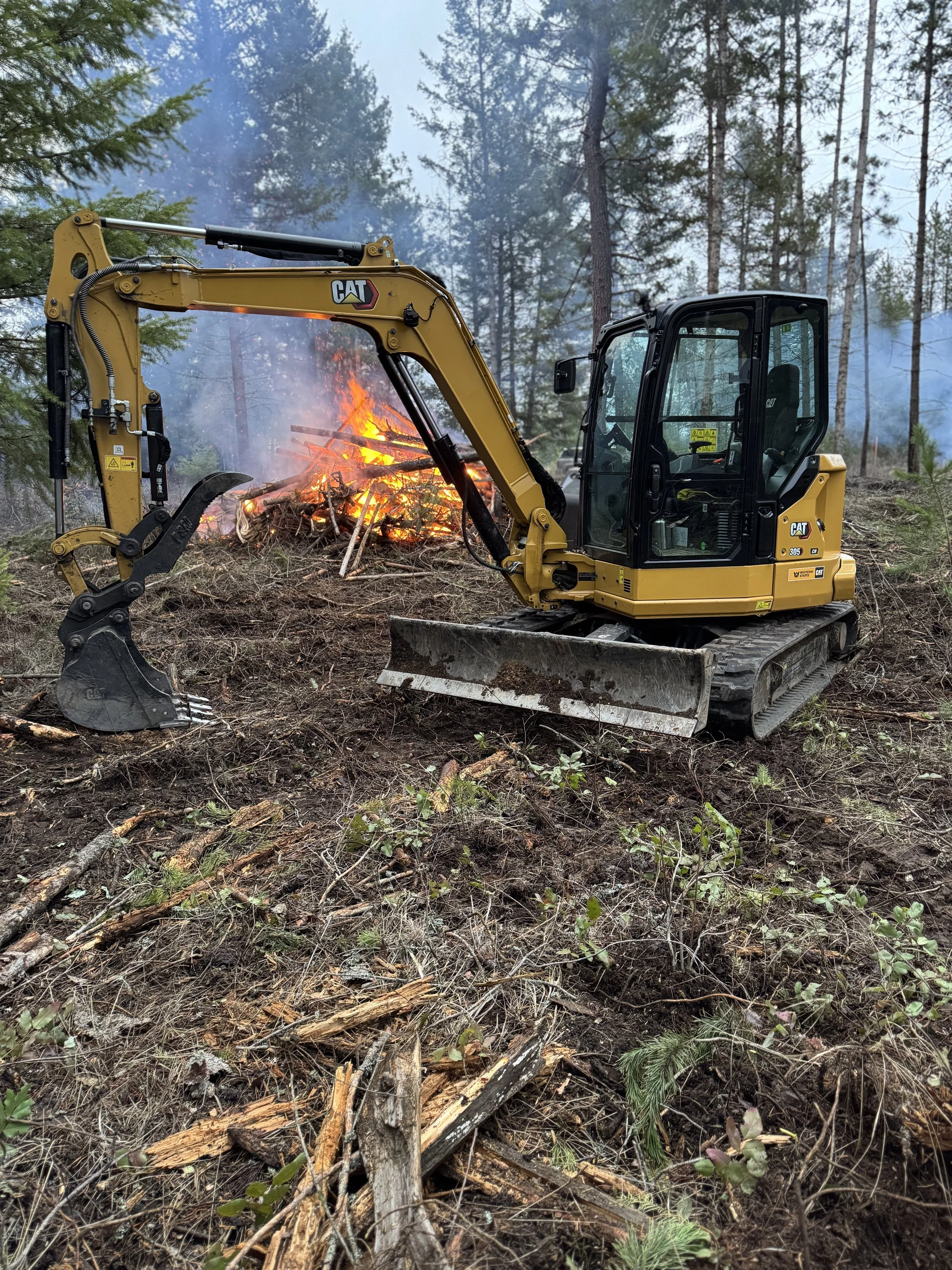 Small yellow CAT excavator is clearing debris on a hillside next to a controlled fire with flames and smoke in a forested area.