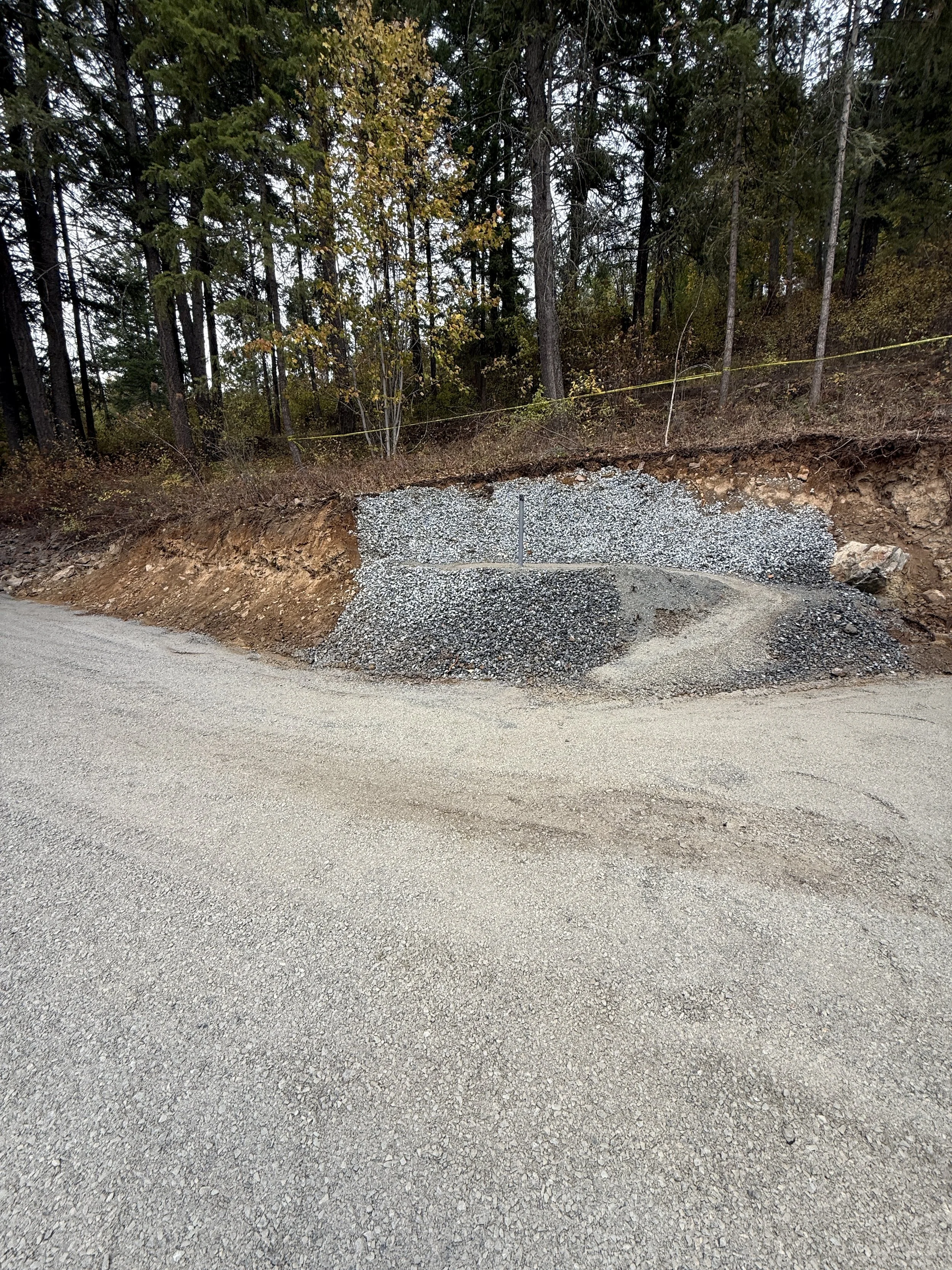 A gravel road edge with a section of exposed dirt and rocks at the roadside, backed by a forest with tall trees and autumn foliage.