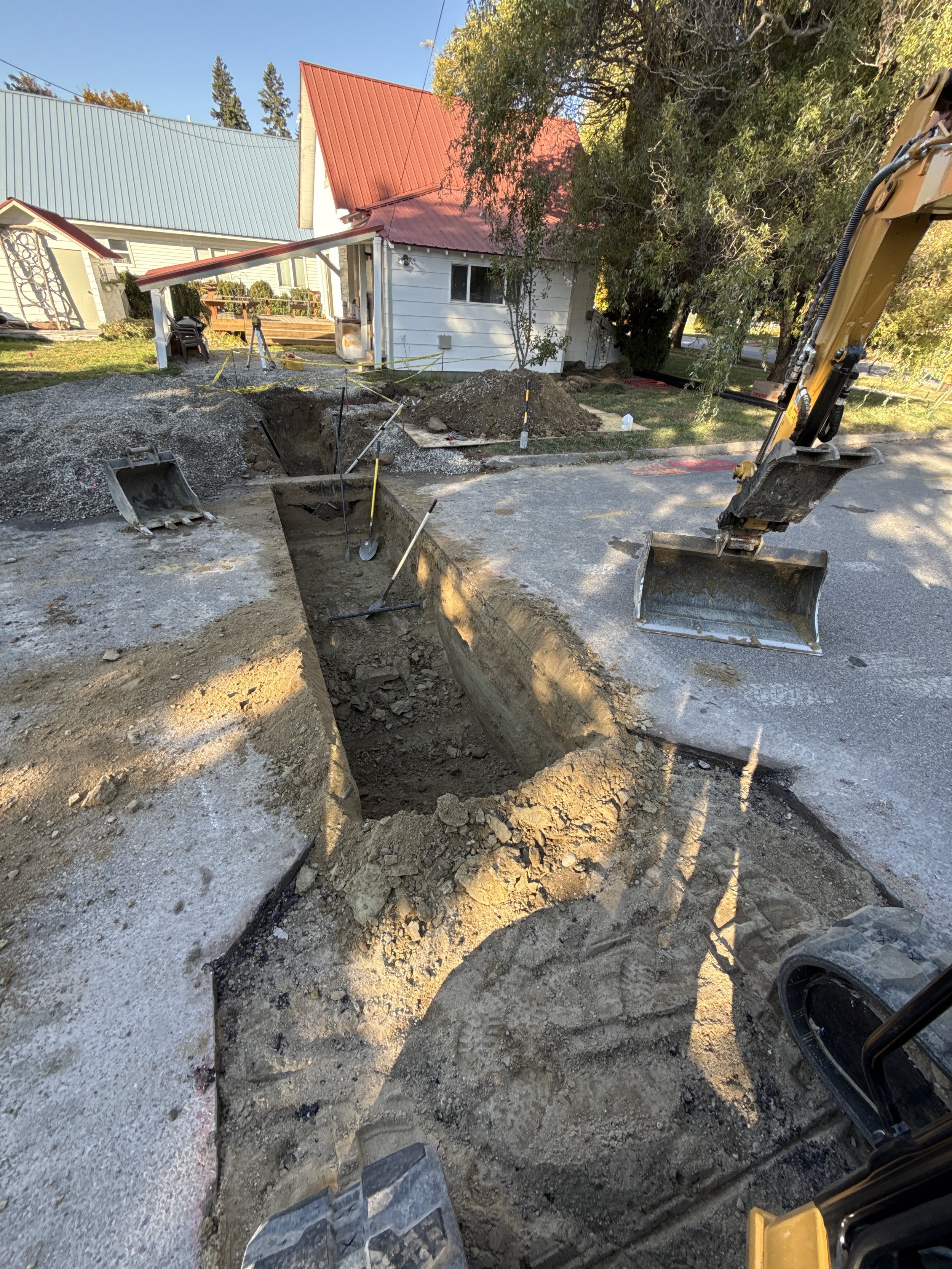 Construction site with a deep trench in the street, a small excavator, and construction tools. In the background, there are houses, trees, and clear sky.