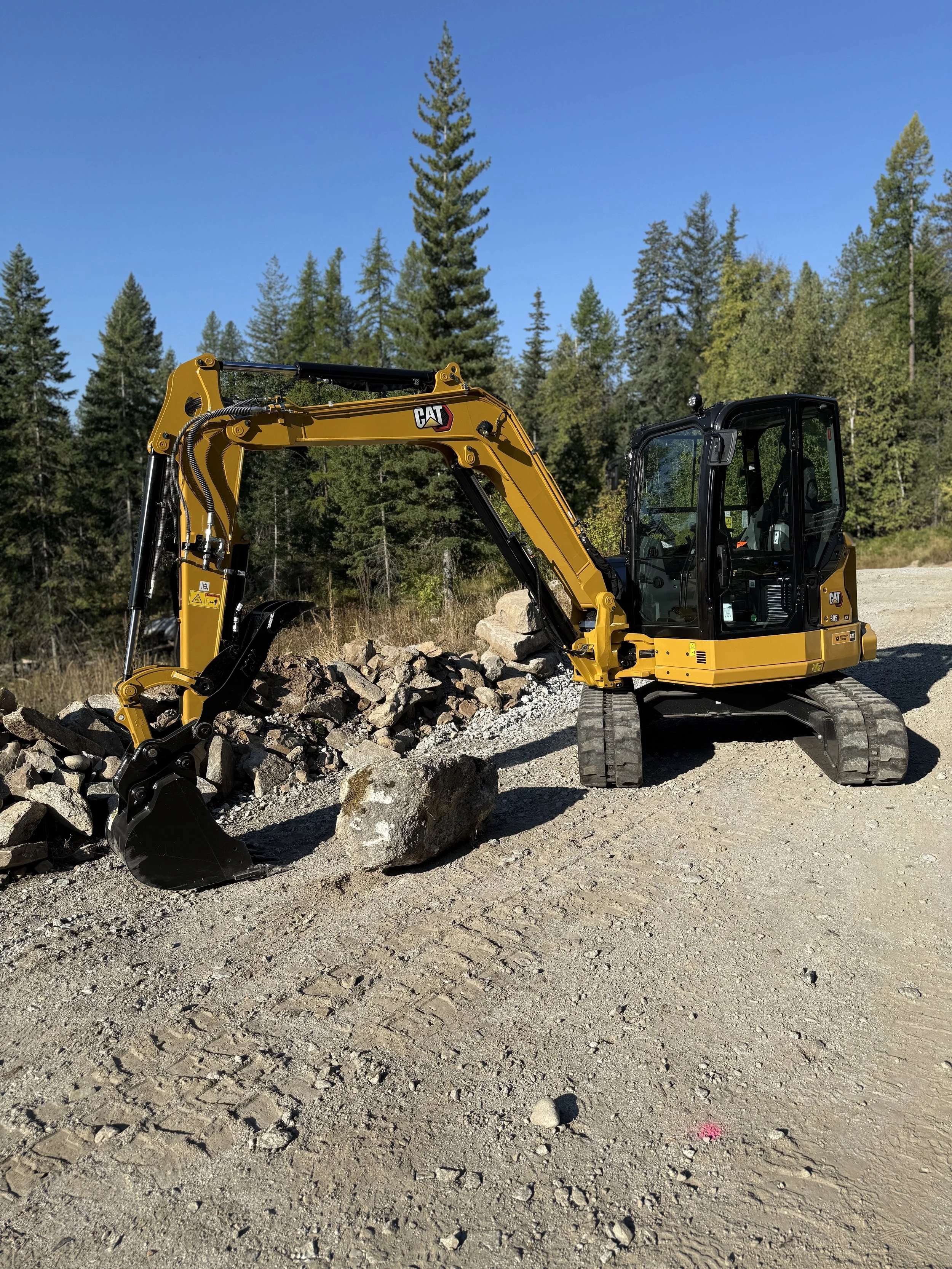 A yellow CAT mini excavator working on a rocky construction site with trees and blue sky in the background.