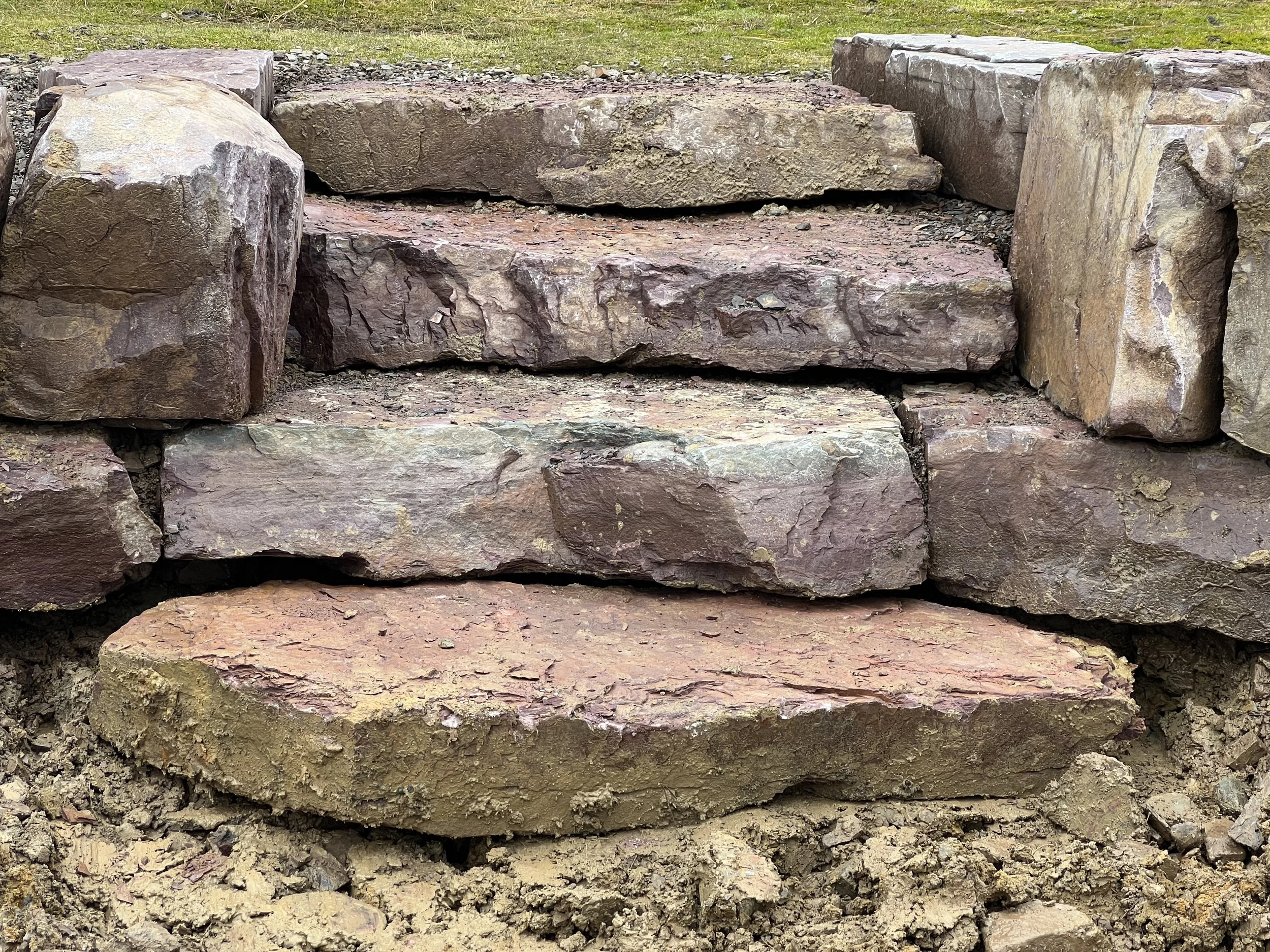 Close-up of a stone staircase made of large, uneven, reddish-brown rocks with soil at the base.