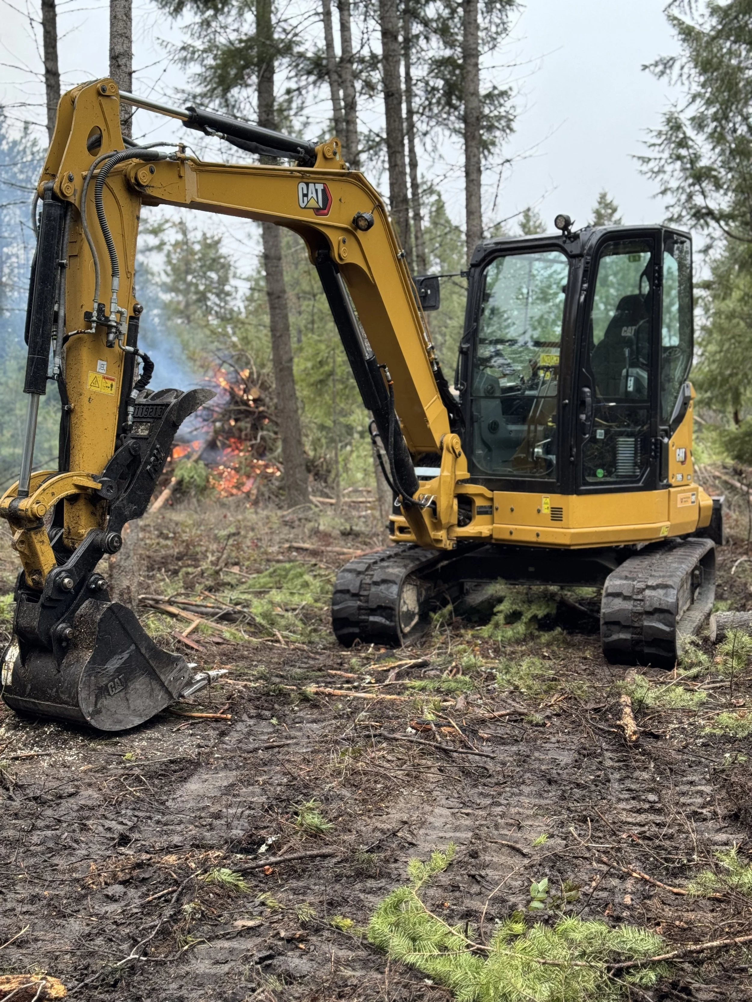 A yellow Caterpillar excavator working in a forested area with trees and a fire burning in the background.