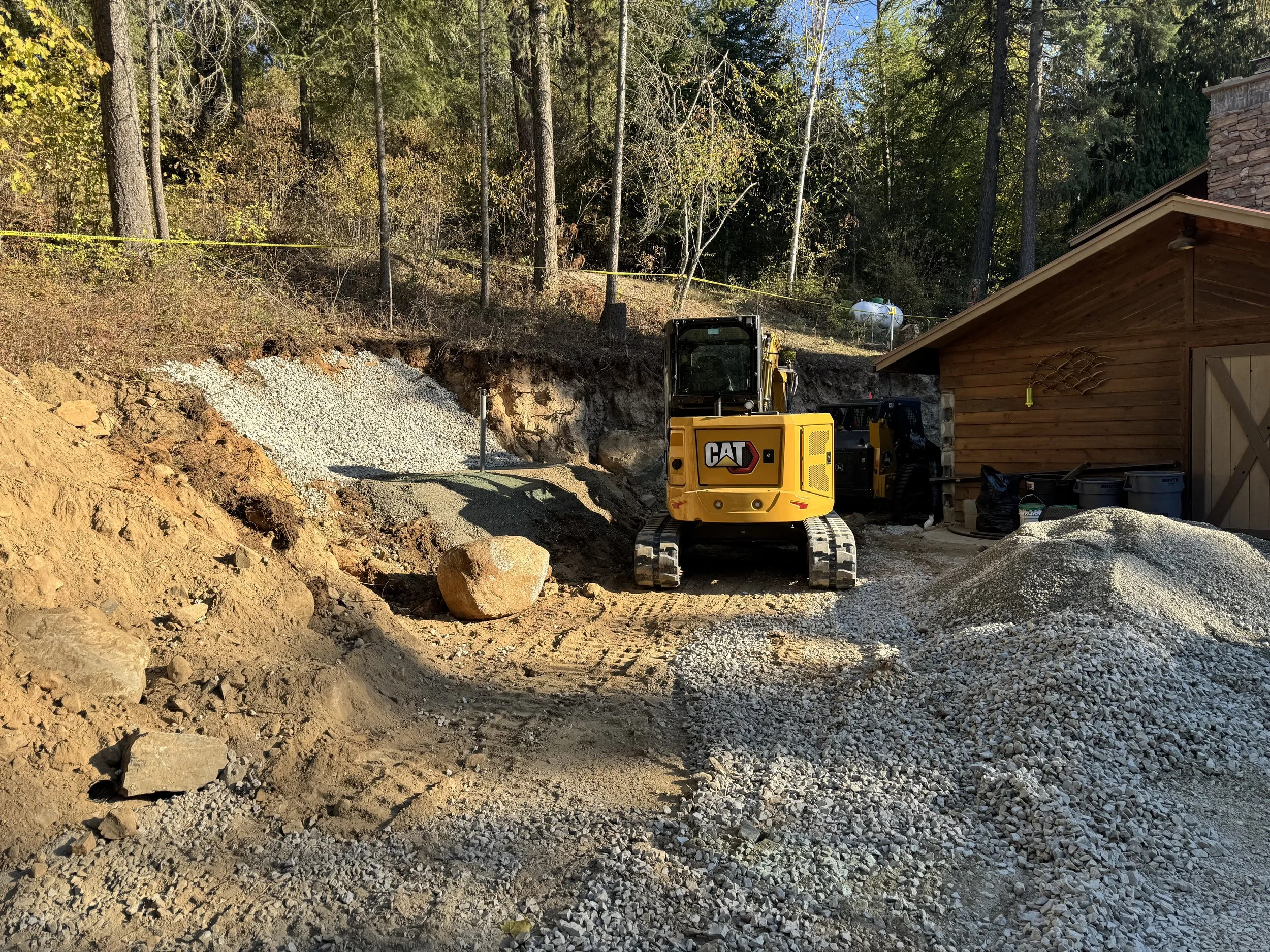 A construction site with a small Caterpillar excavator, piles of gravel and dirt, a wooden shed, and a wooded background.