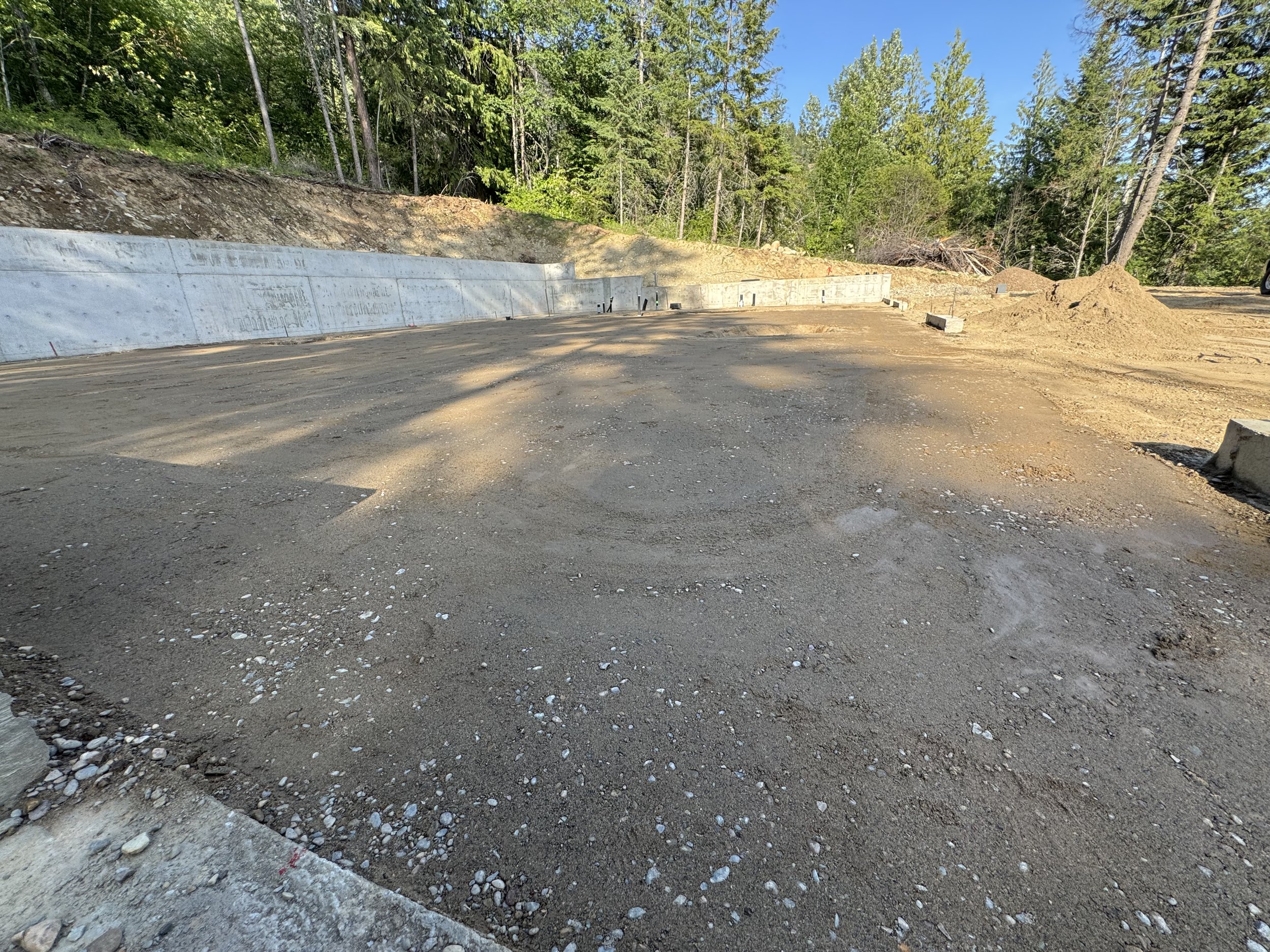 Construction site with an uncovered dirt ground, concrete barriers, and trees in the background under a clear blue sky.