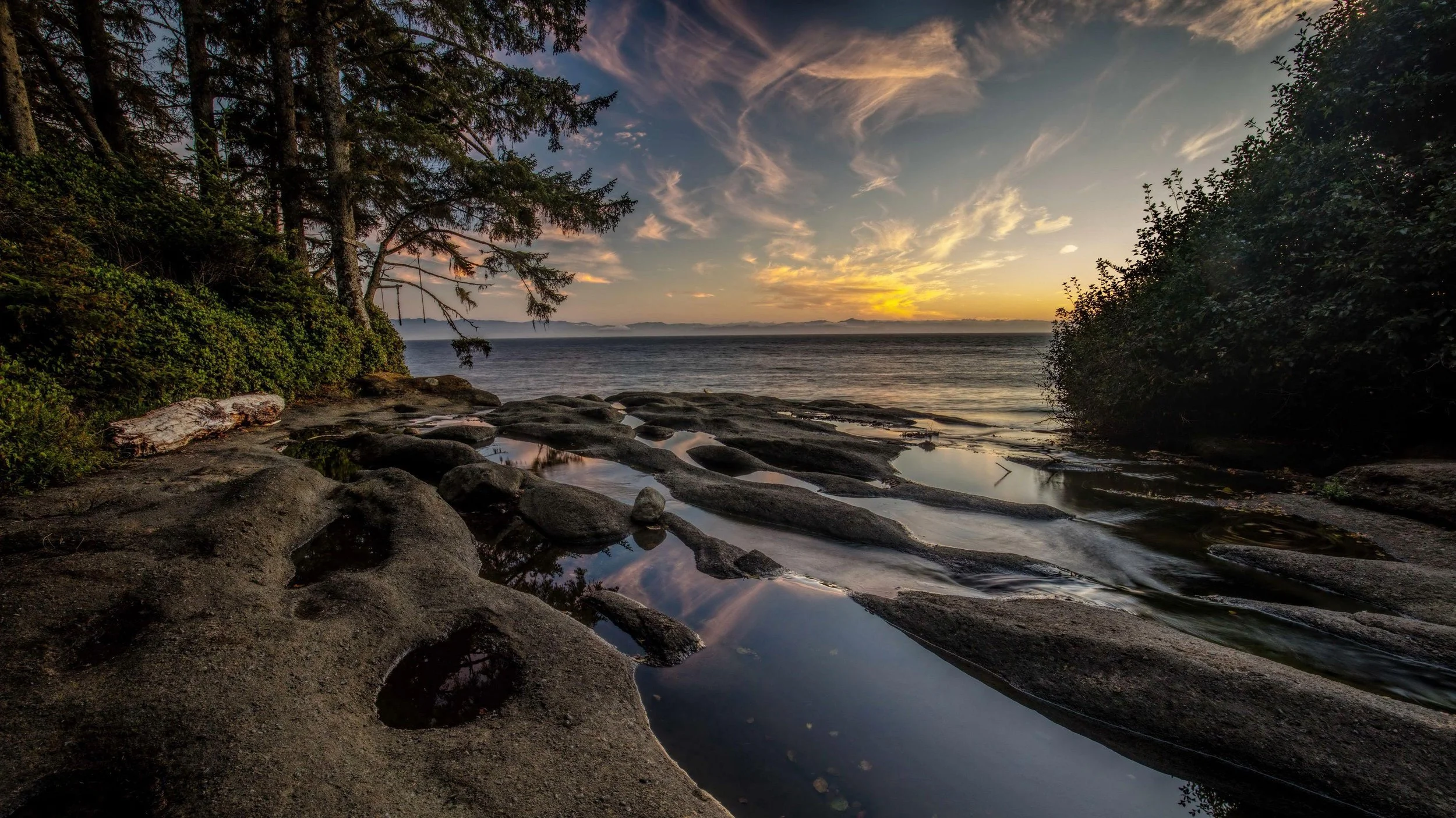 Sunset over a rocky shoreline with water reflecting the sky, trees and shrubbery on both sides.