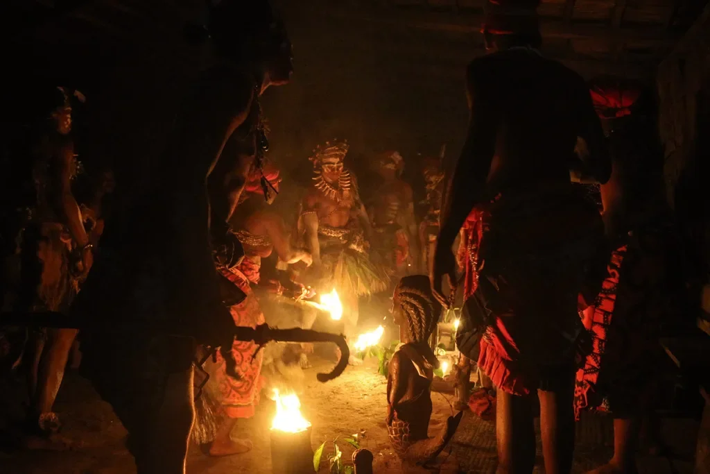 Indigenous people performing a traditional ceremony in a dimly lit space with fire and cultural attire.