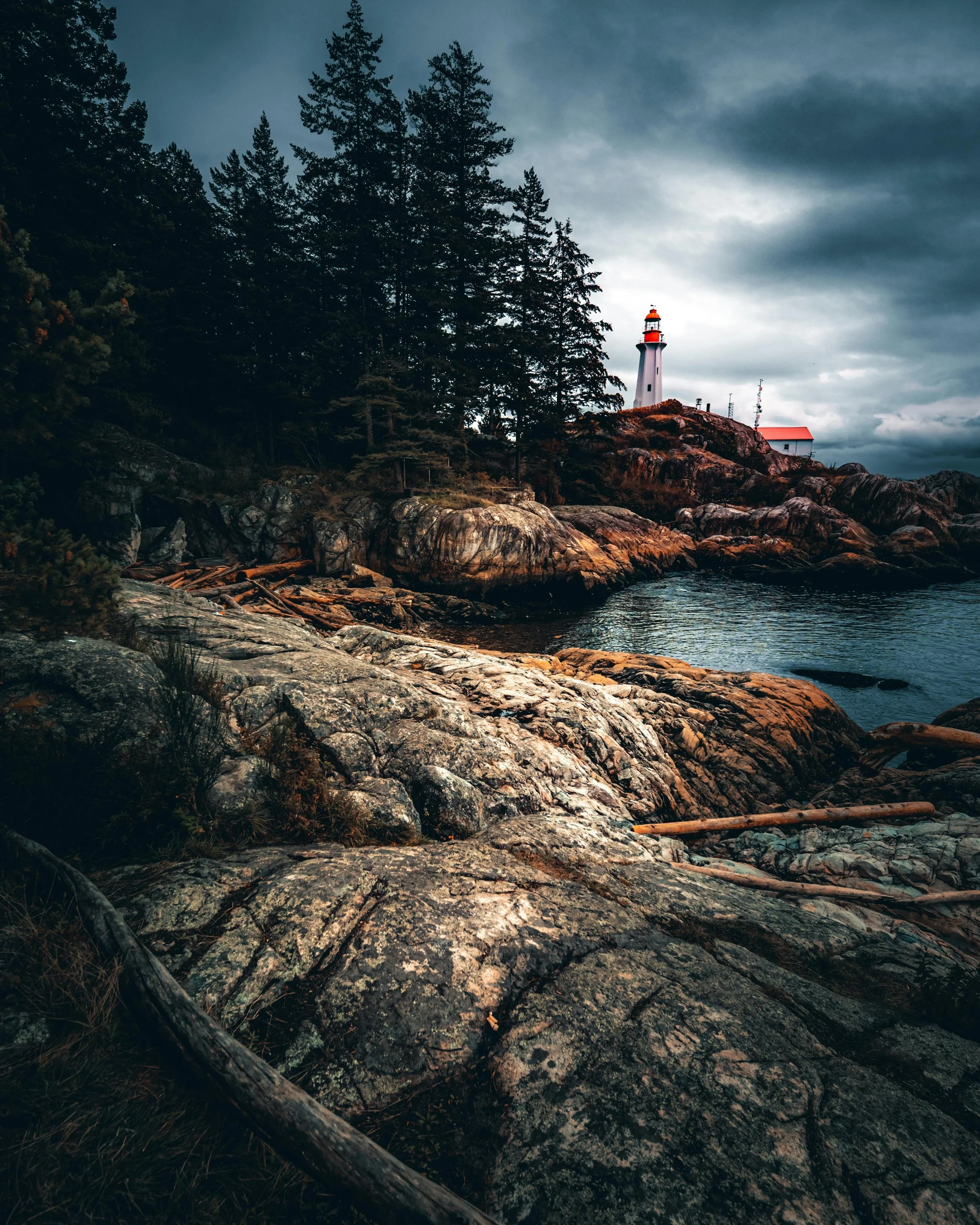 A lighthouse stands on a rocky coastline in vancouver BC with tall evergreen trees nearby, under a cloudy sky at dusk.