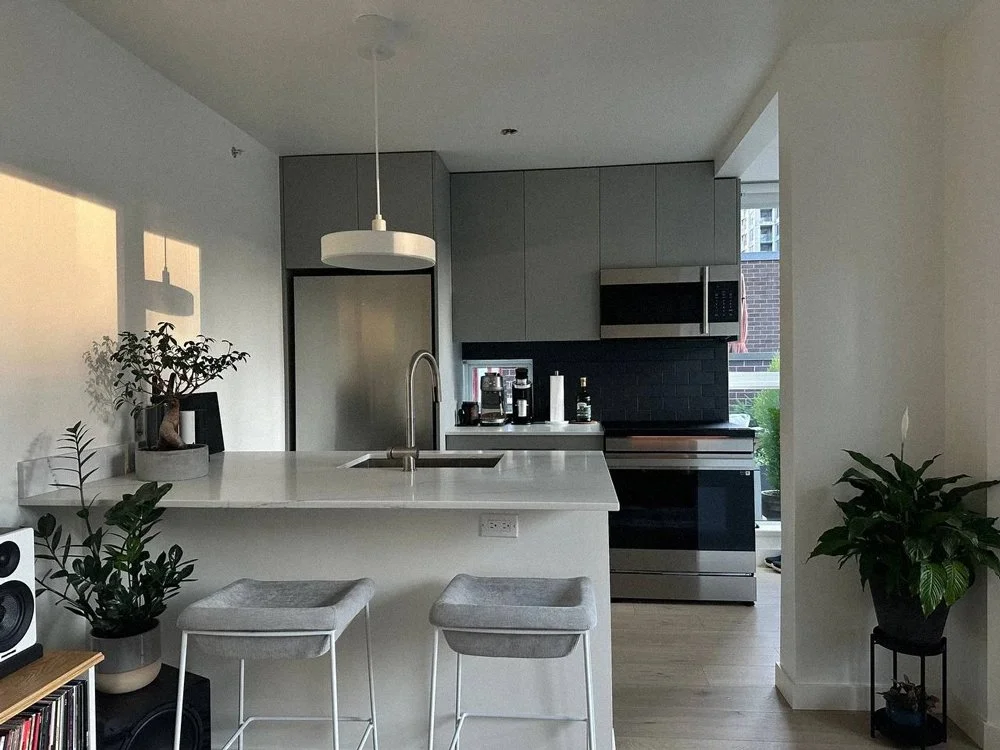 Modern kitchen with gray cabinets, stainless steel appliances, white countertop island with a sink, two gray bar stools, potted plants, and a coffee maker.
