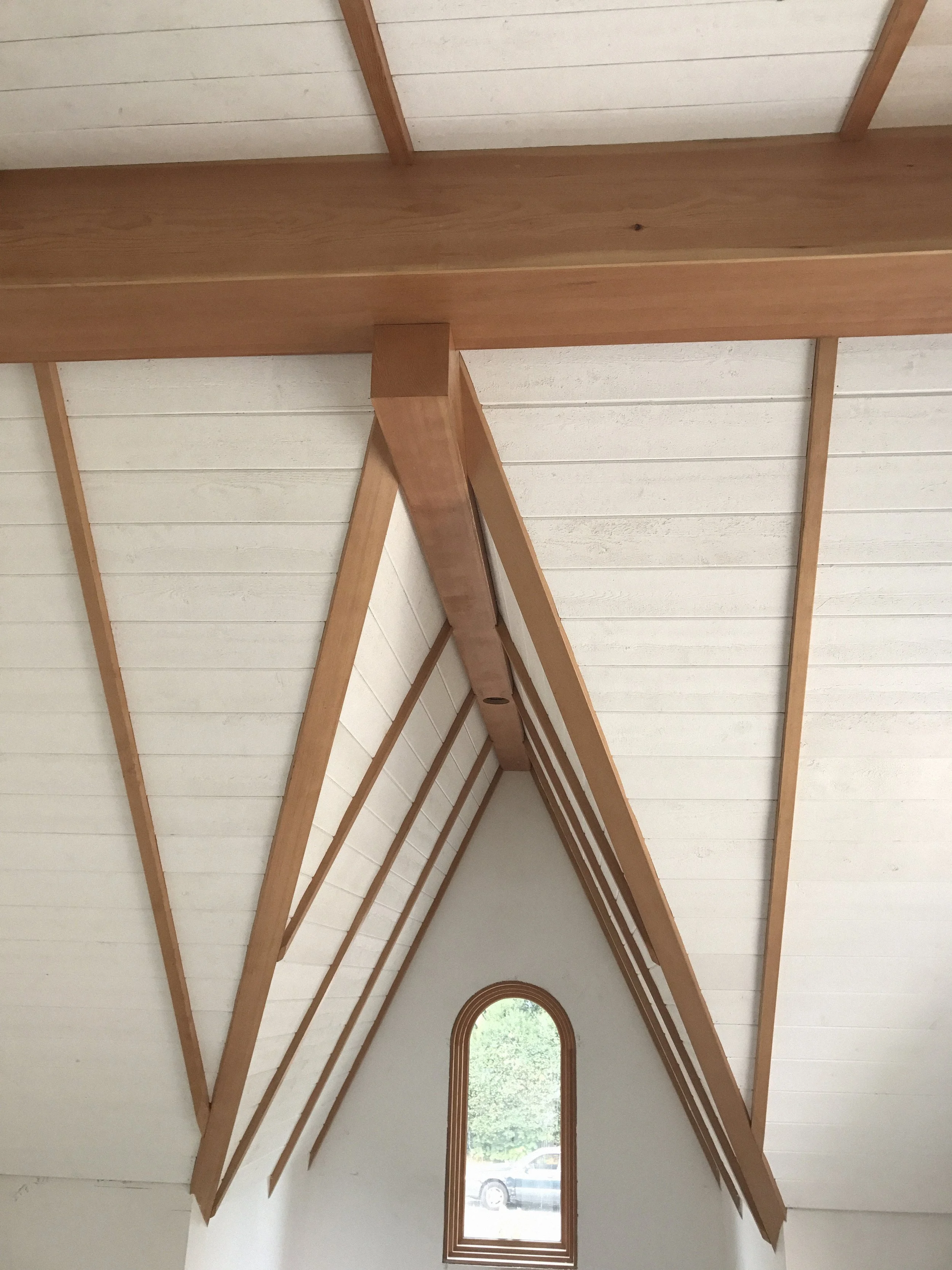 Interior of a building with a vaulted ceiling featuring exposed wooden beams and white wooden panels, and a small arched window letting in natural light.