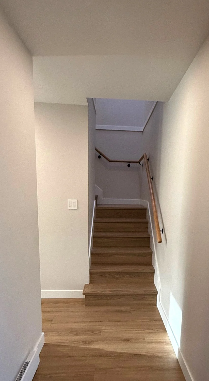 Interior view of a staircase with wooden steps, handrail, and walls painted in light colors, leading to an upper level.