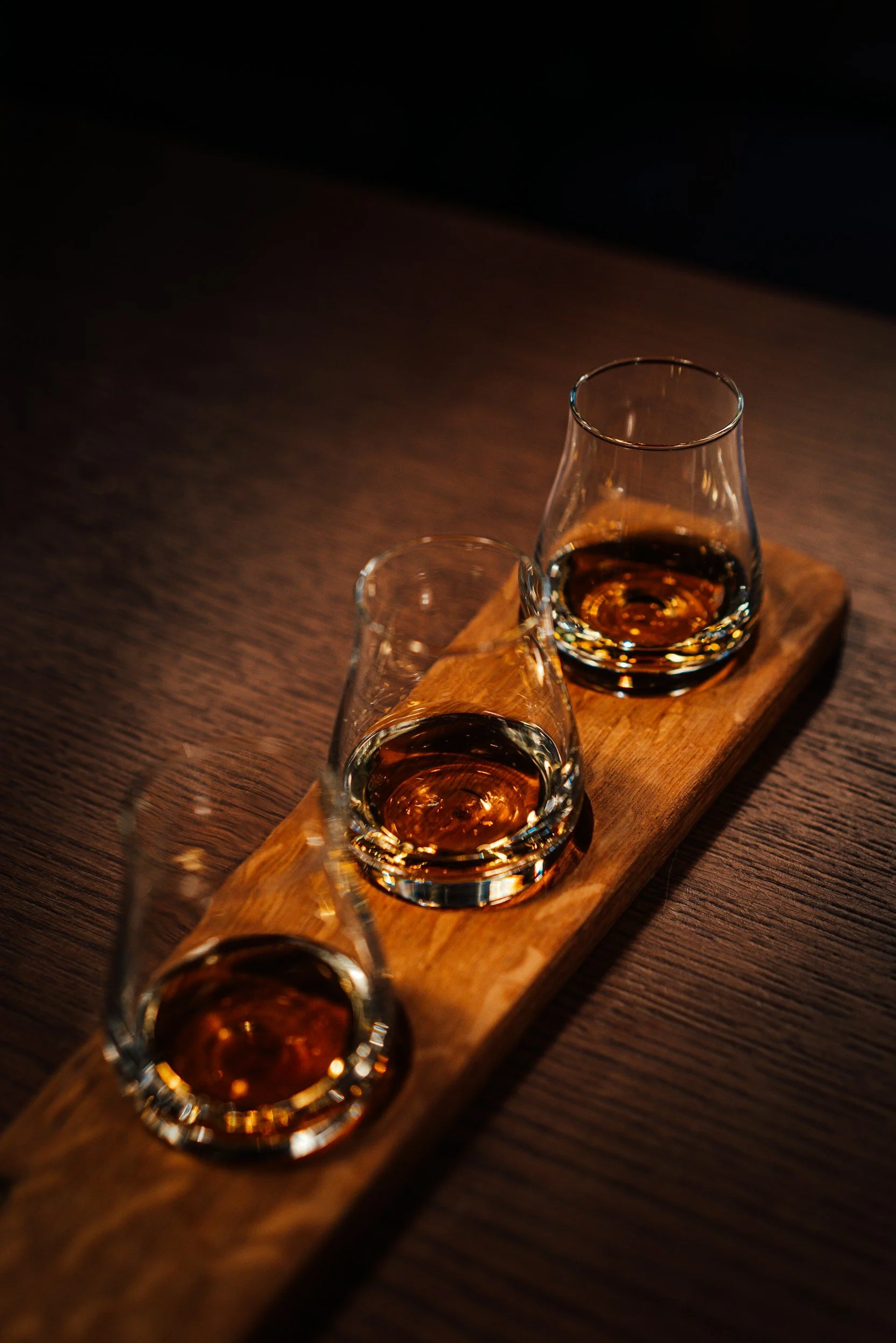 Three glasses of whiskey on a wooden tray with dark background.
