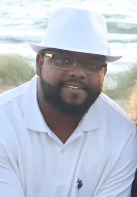 Man wearing a white hat, glasses, and a white polo shirt, smiling at the camera, with the beach and ocean in the background.
